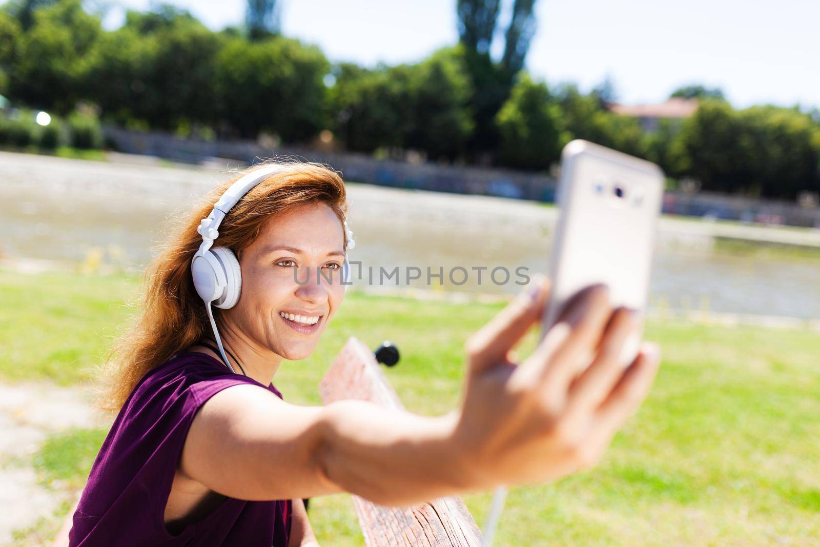 brunette girl with headphones, taking a selfie while sitting on a bench by kokimk