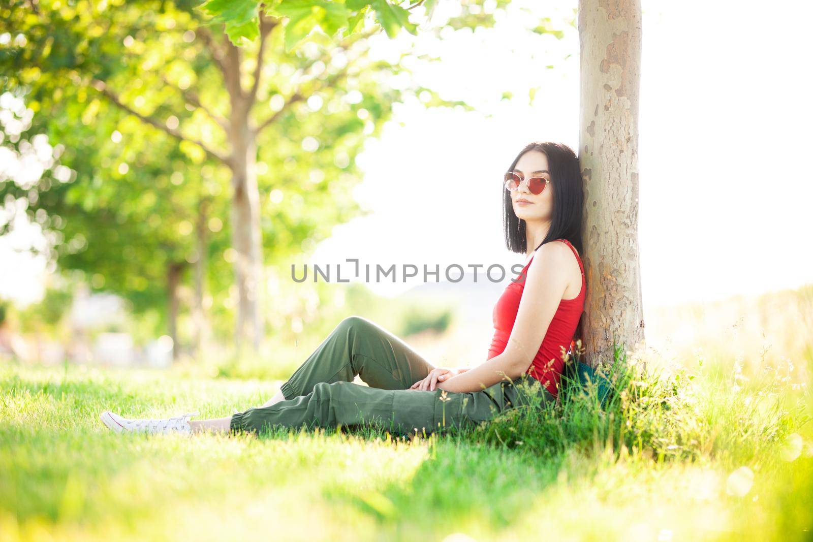 girl with dark brown hair and red sunglasses posing under a tree