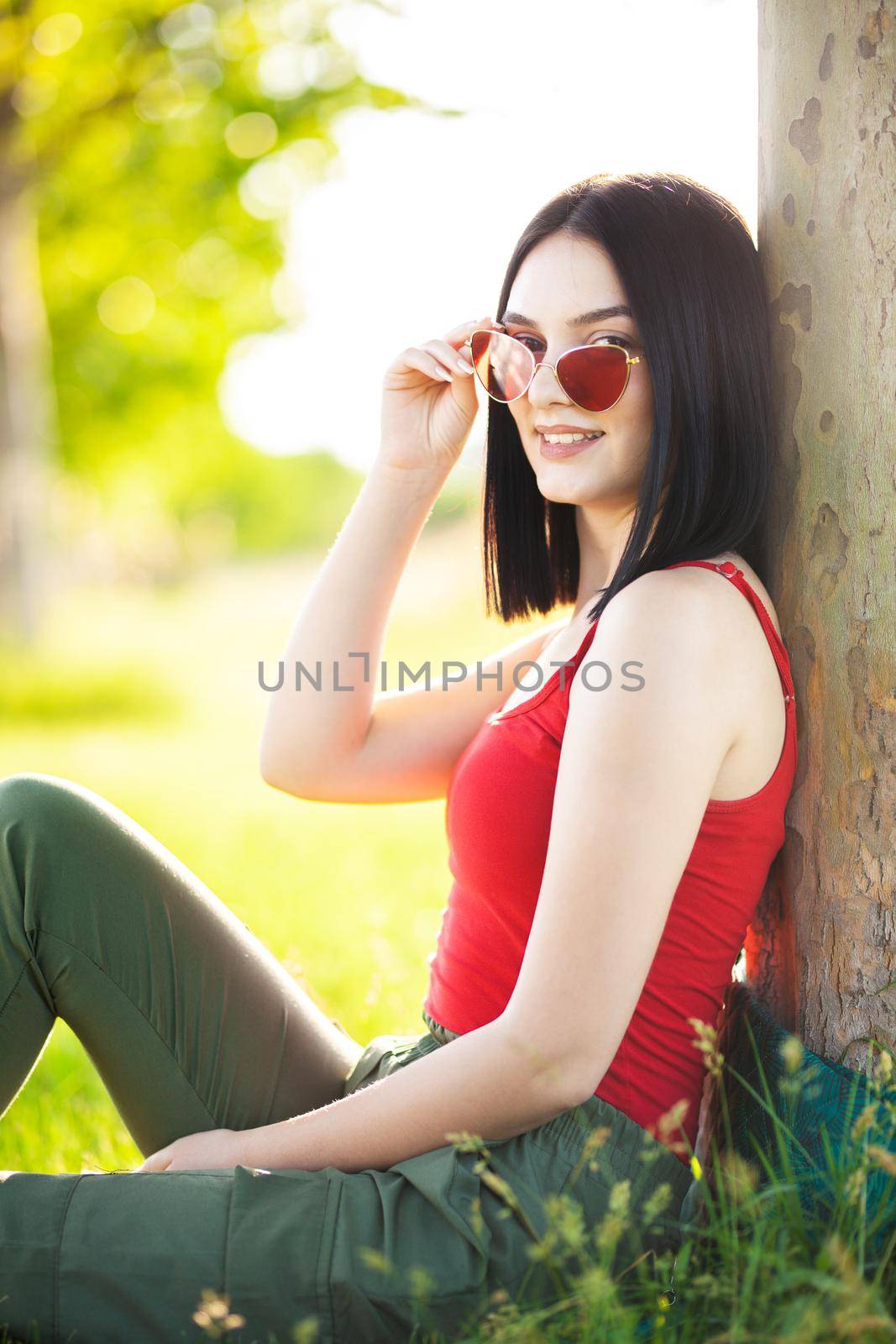 girl with dark brown hair and red sunglasses posing under a tree