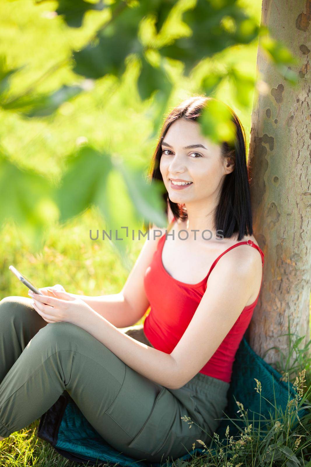 girl with dark brown hair using smartphone while sitting under a tree