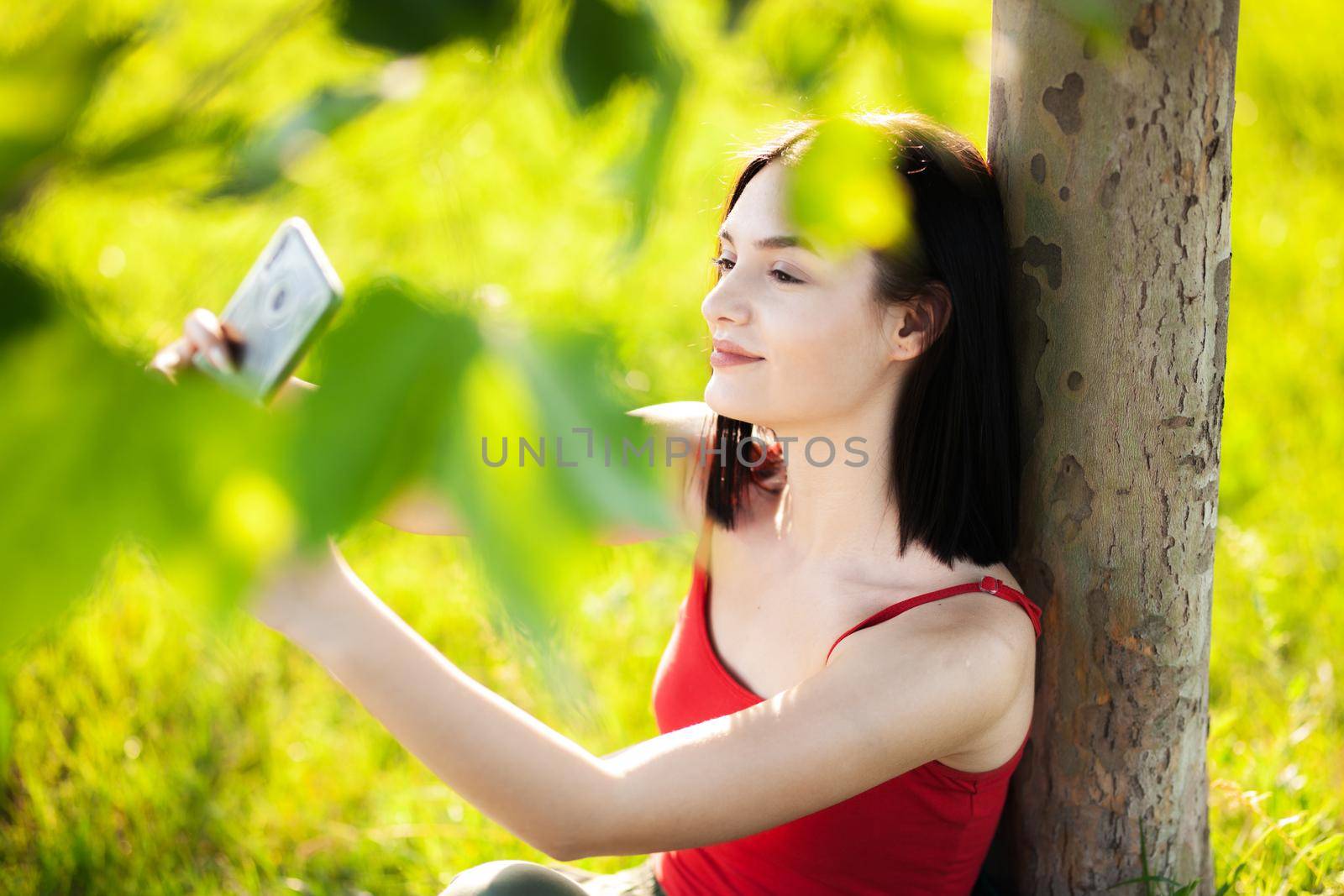 girl with dark brown hair using smartphone taking selfy under a tree