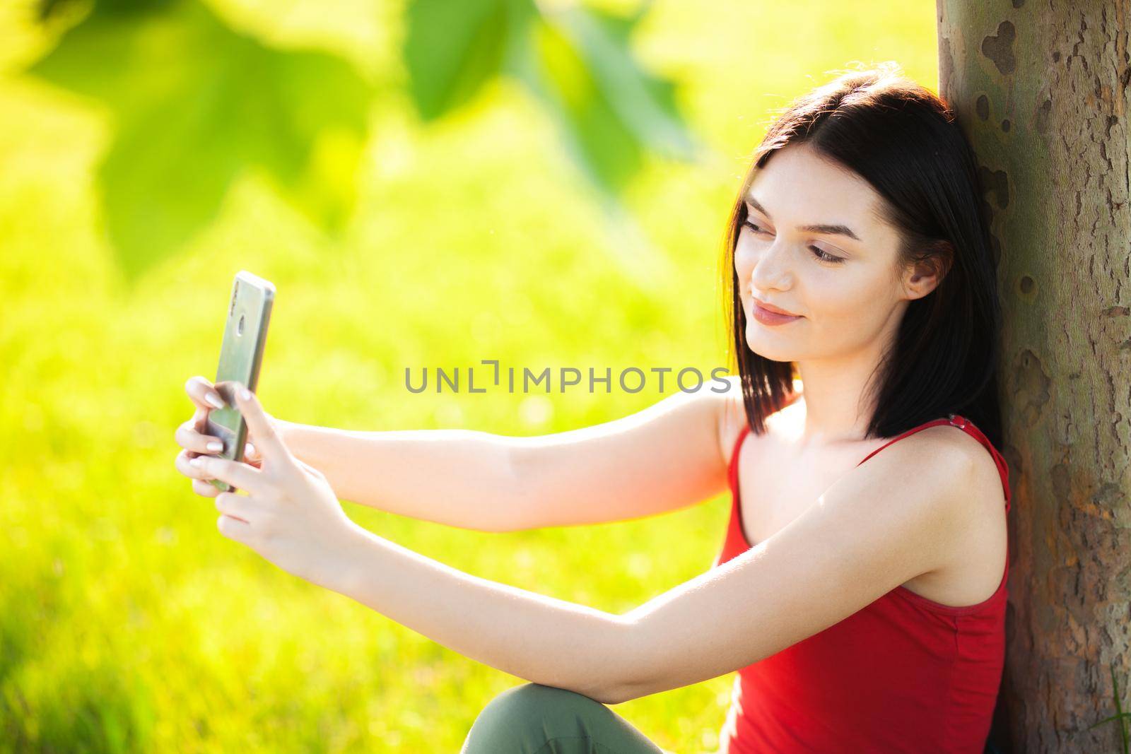 girl with dark brown hair using smartphone taking selfy under a tree