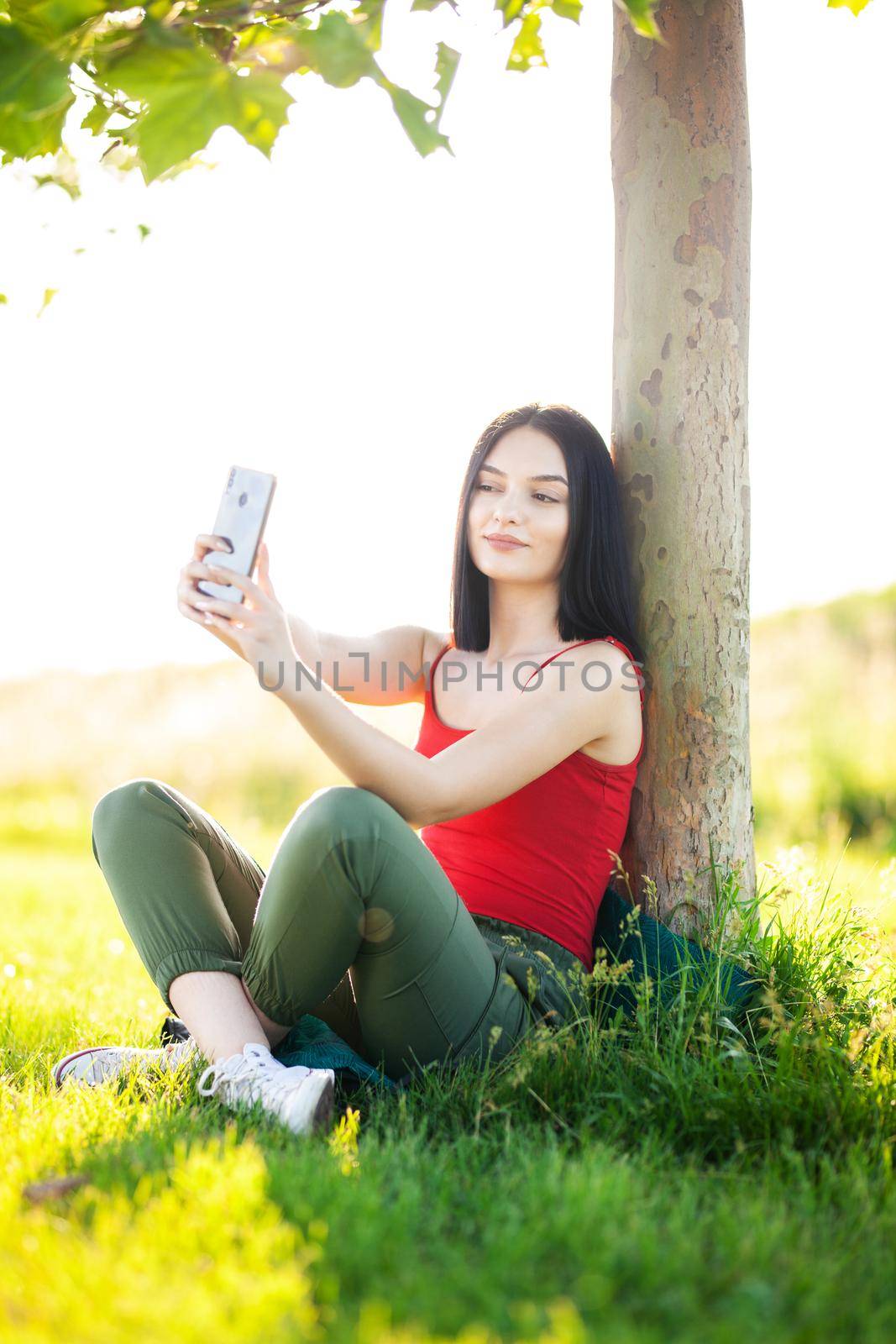 girl with dark brown hair using smartphone taking selfy under a tree