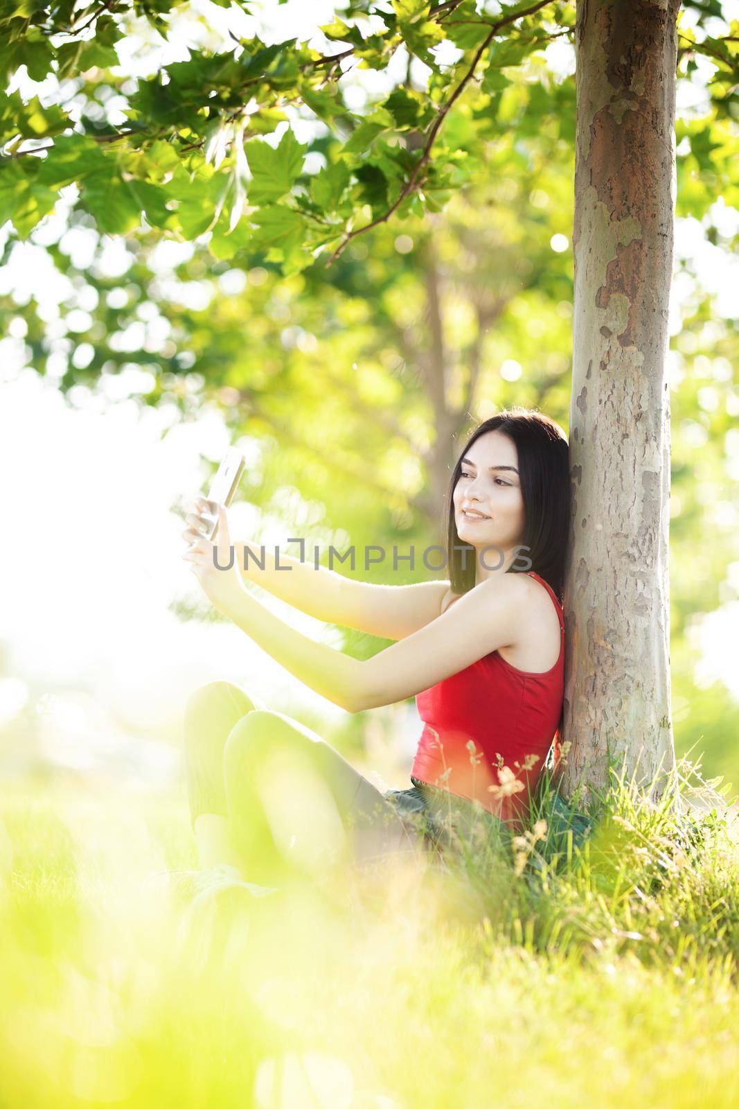 girl with dark brown hair using smartphone taking selfy under a tree by kokimk