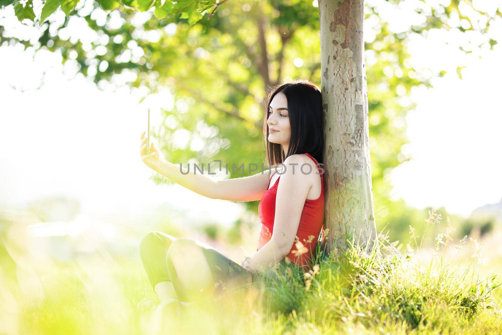 girl with dark brown hair using smartphone taking selfy under a tree