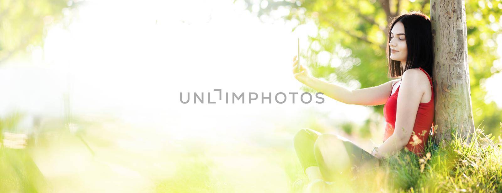 girl with dark brown hair using smartphone taking selfy under a tree