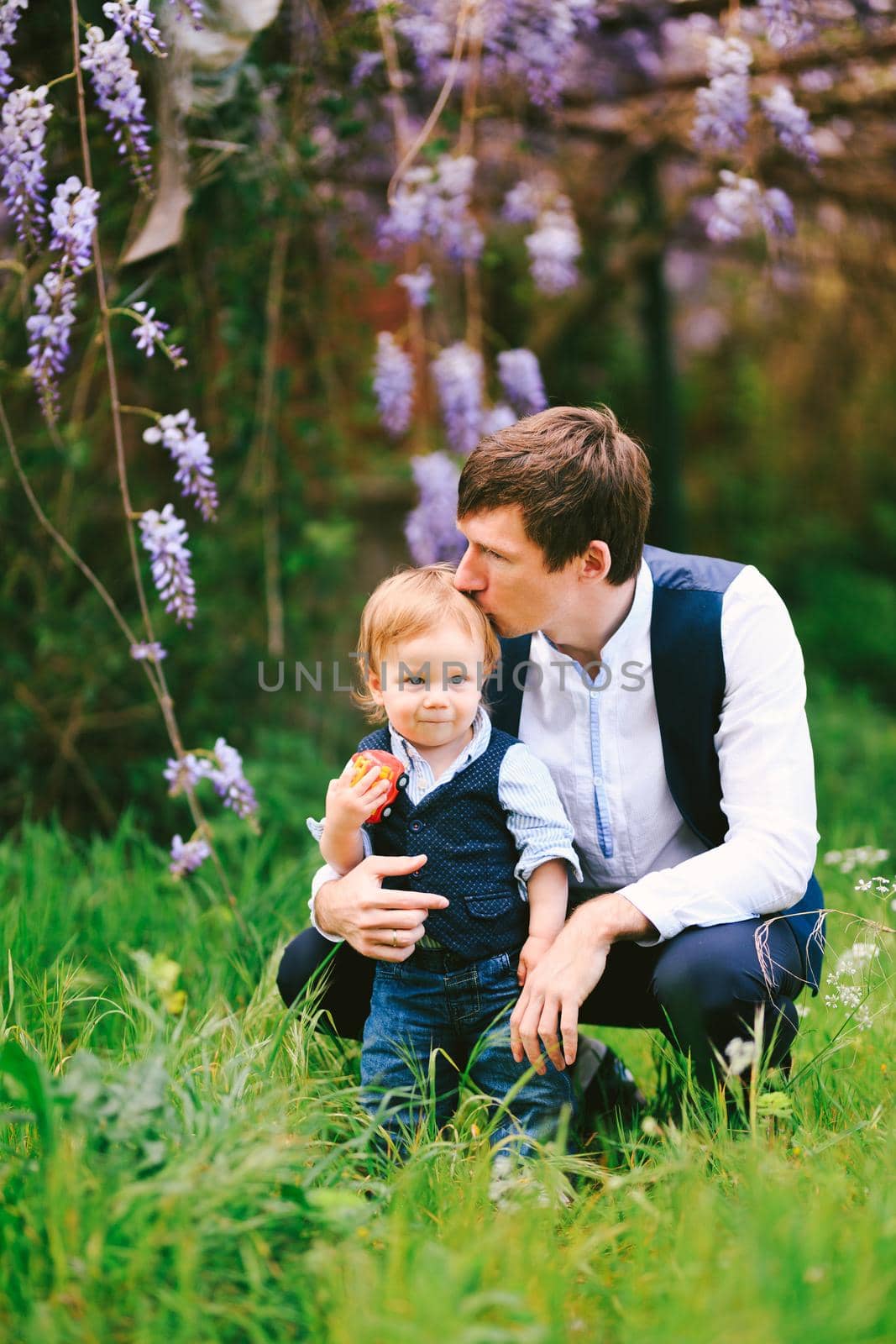 Father is kissing his baby boy on top of the head with a wysteria blooming in the background by Nadtochiy