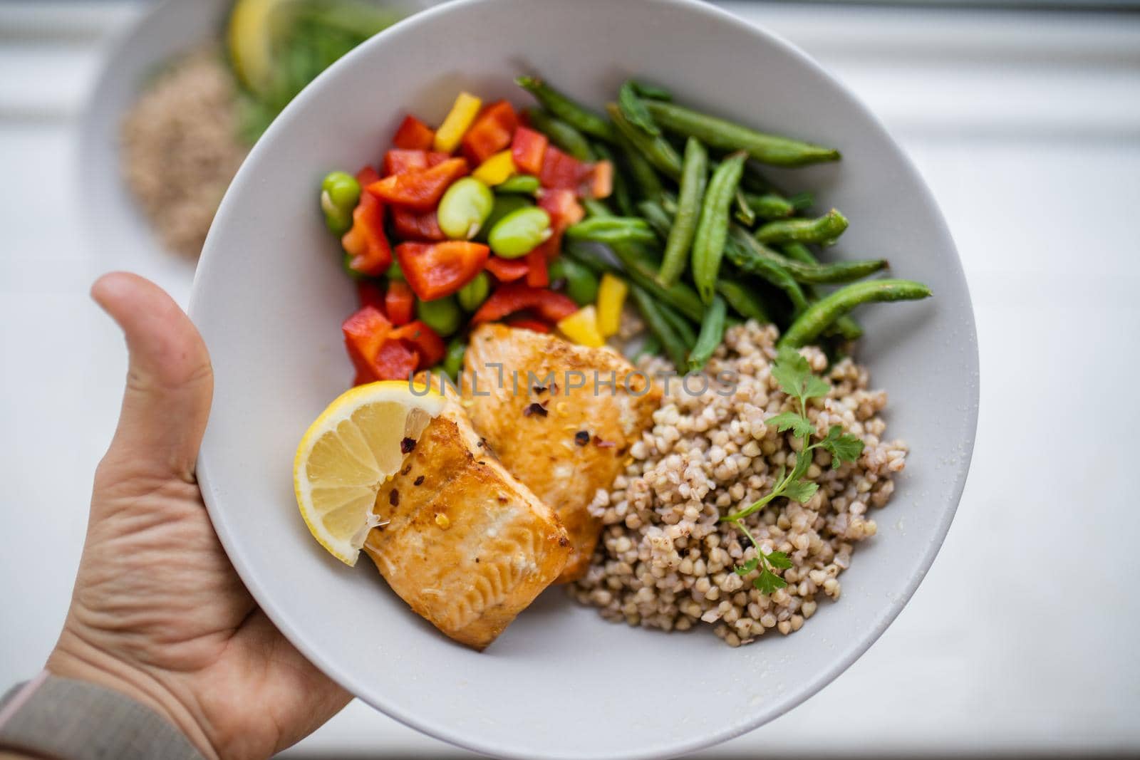 Hand holding salmon and buckwheat dish with green beans, broad beans, and tomato slices. Nutritious dish with vegetables and fish on white plate. Healthy balanced diet
