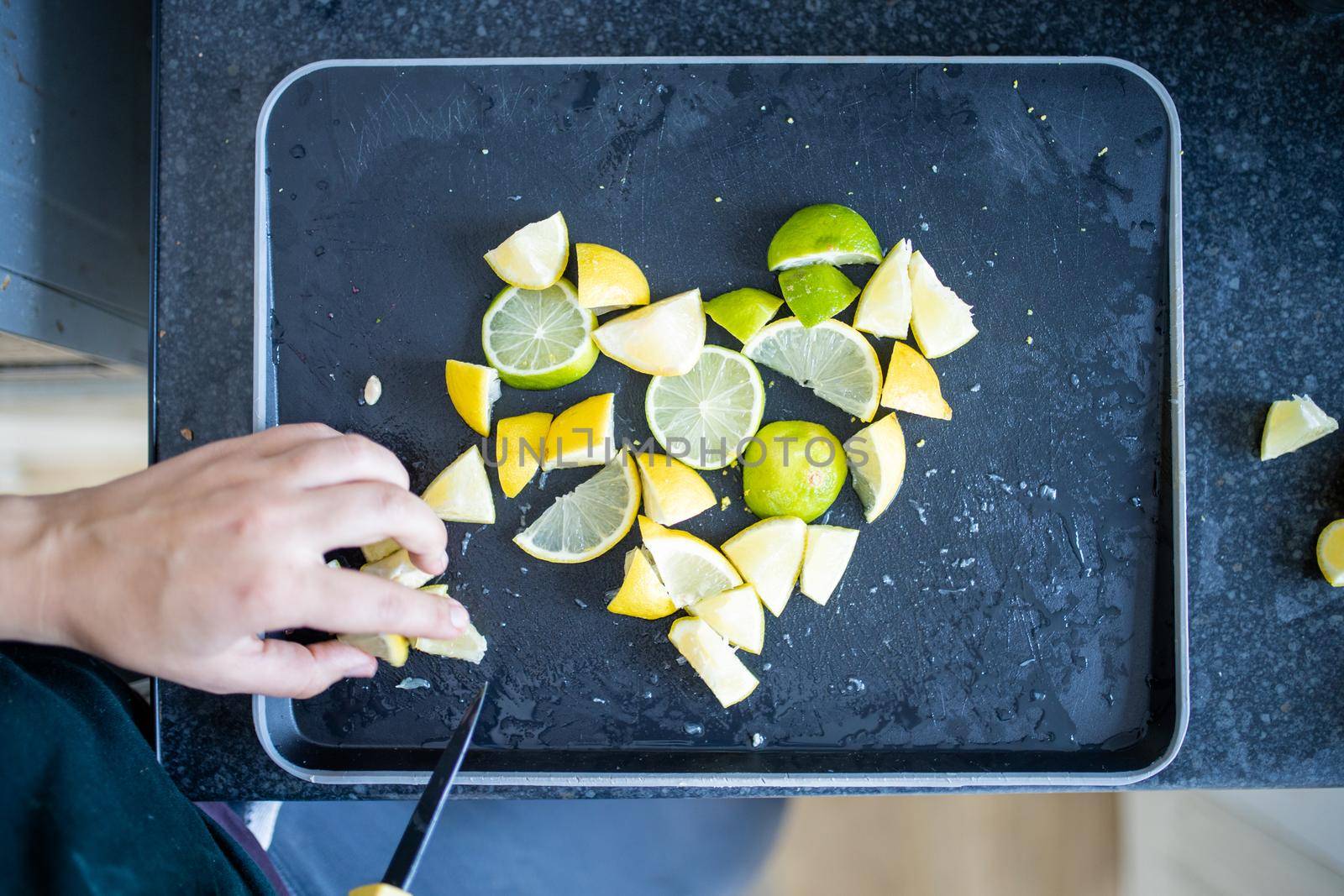 Top view of female hands carefully slicing lemons and limes on black tray. Woman cutting citric fruit into small pieces forming a heart. Citrus drink preparation