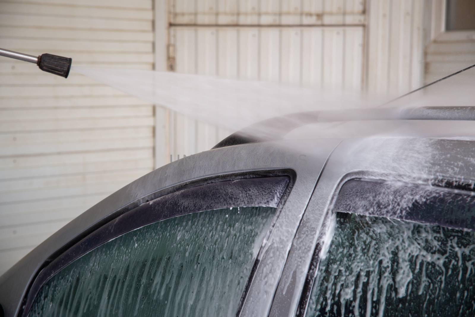 Indoor self service car wash process - close-up with selective focus and background blur. High pressure stream of water spraying on silver car's roof.