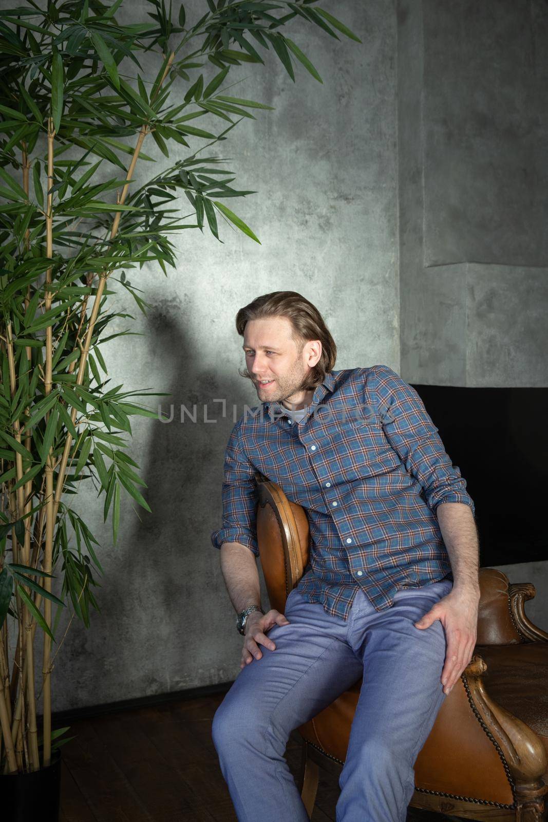 The handsome stylish young man sits on an expensive leather armchair relaxed, long curly hair, he is dressed in a blue shirt and trousers, he is not looking at the camera. High quality photo