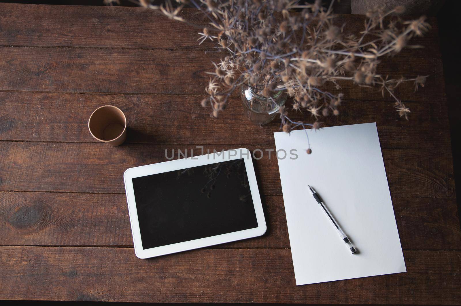 Modern office workplace with tablet pc on the table. On the table is a tablet computer with a sheet of white paper, dried flowers in a glass and a disposable cup of coffee.