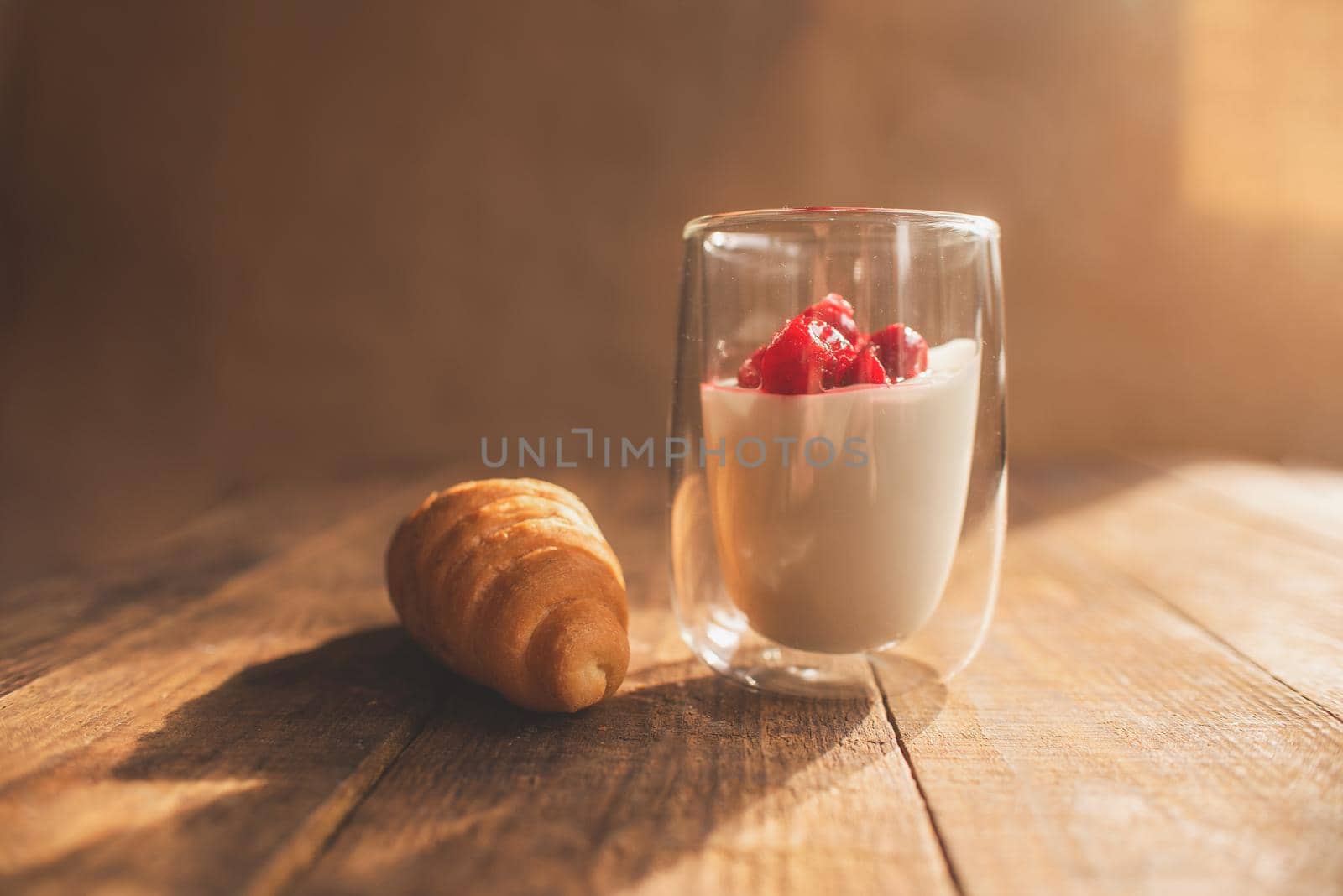double glass with yogurt and berries and croissant on a wooden table where the bright sun is shining