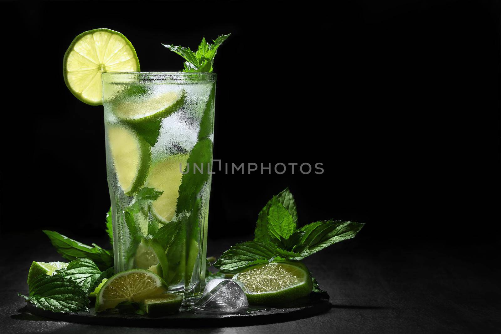 Fresh homemade mojito cocktail in a tall glass with lime, mint and ice on a black background, copy space by galsand