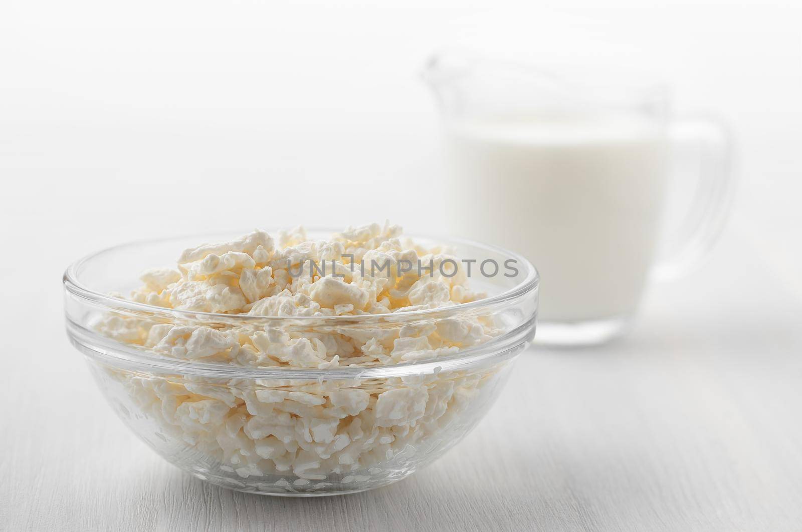 Fresh homemade farm cottage cheese in glass bowl and cream on white wooden table.