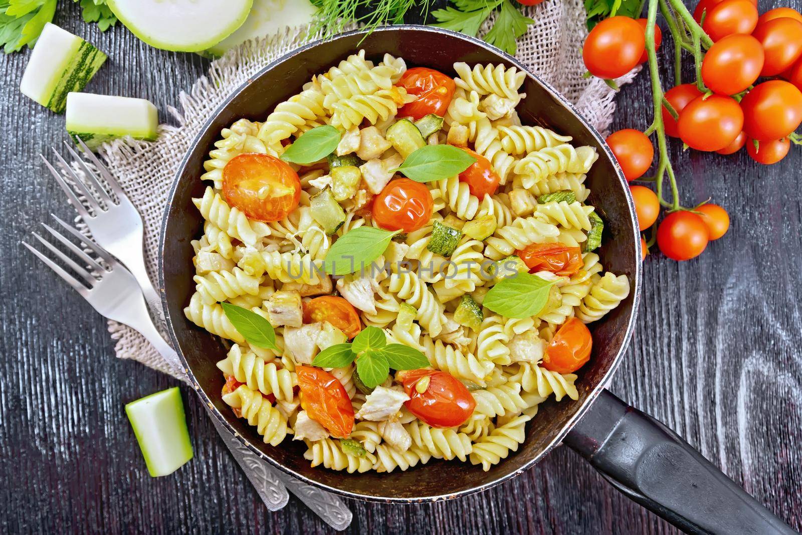 Fusilli with chicken, zucchini and tomatoes in a frying pan on burlap, forks, basil and parsley on black wooden board background from above