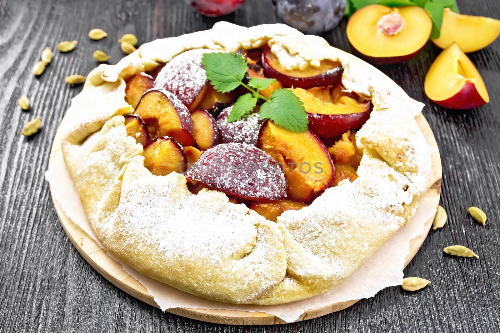 Sweet pie with plum, sugar and cardamom on parchment, sprigs of green mint on a dark wooden board background