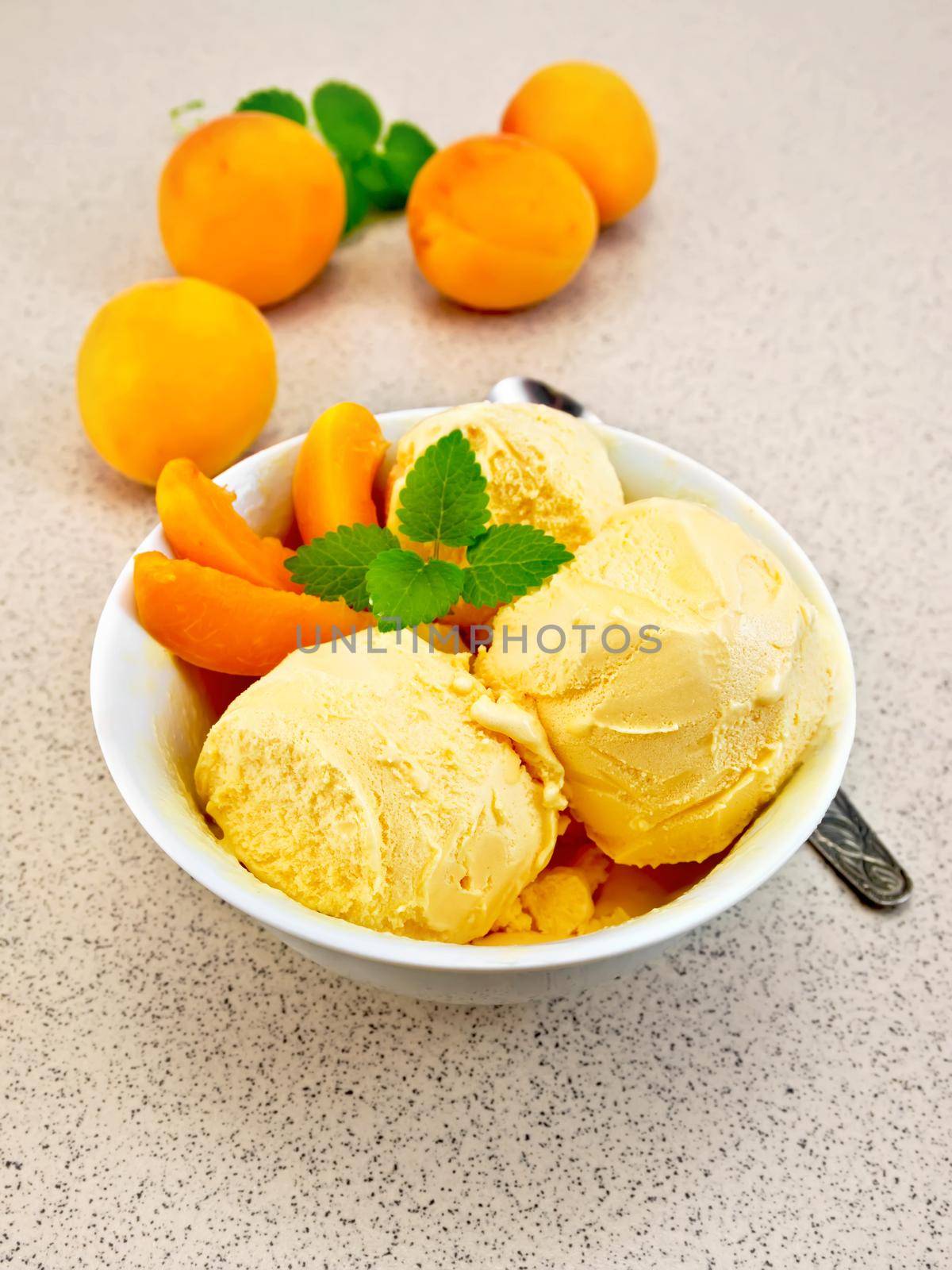 Apricot ice cream in a white bowl with slices of fruit and mint on a background granite table