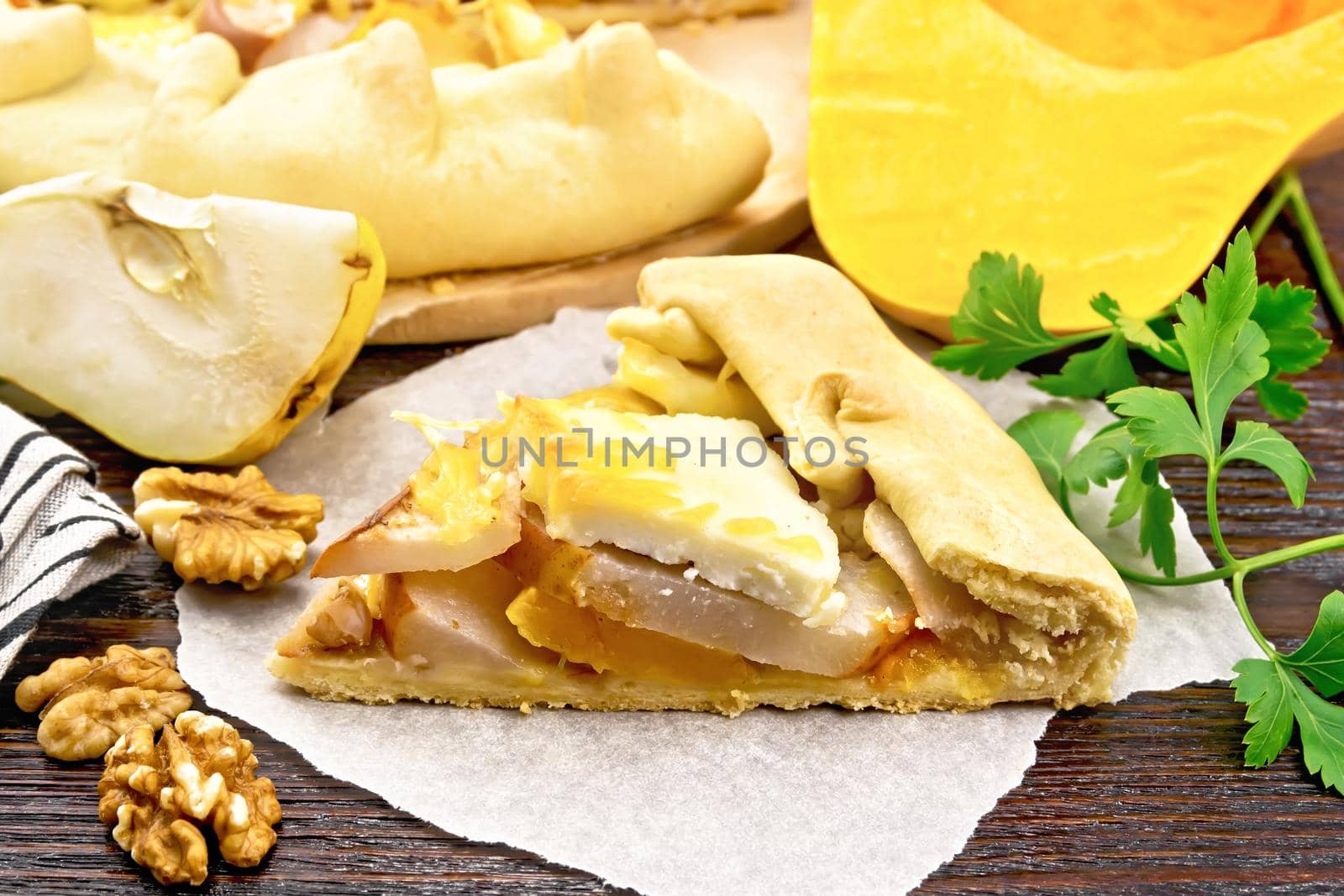 Piece pie of pumpkin, pear, soft cheese and walnuts on a parchment, parsley on a wooden board background