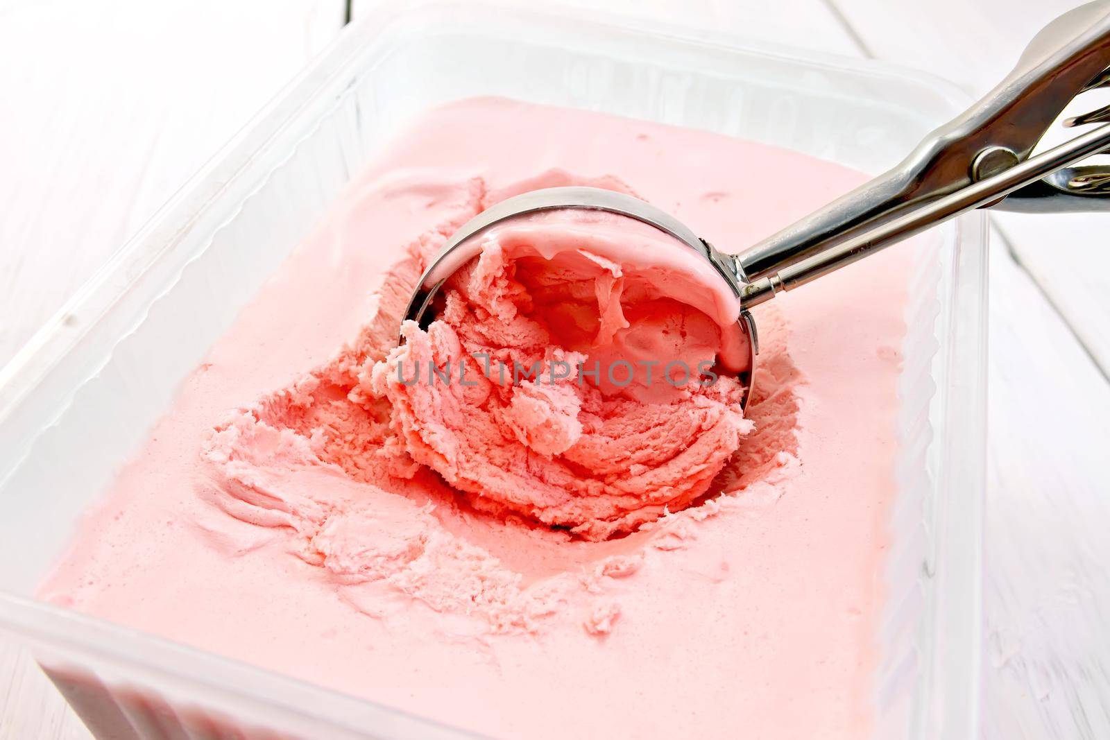 Strawberry ice cream in a plastic tray and a metal scoop on background light wooden boards