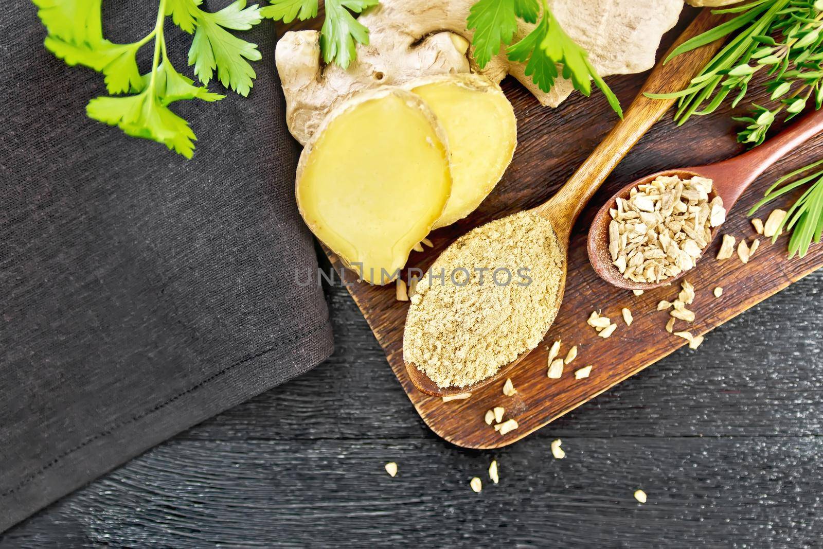 Ground ginger and flakes in two wooden spoons, ginger root, spicy herbs, napkin on a wooden board background from above