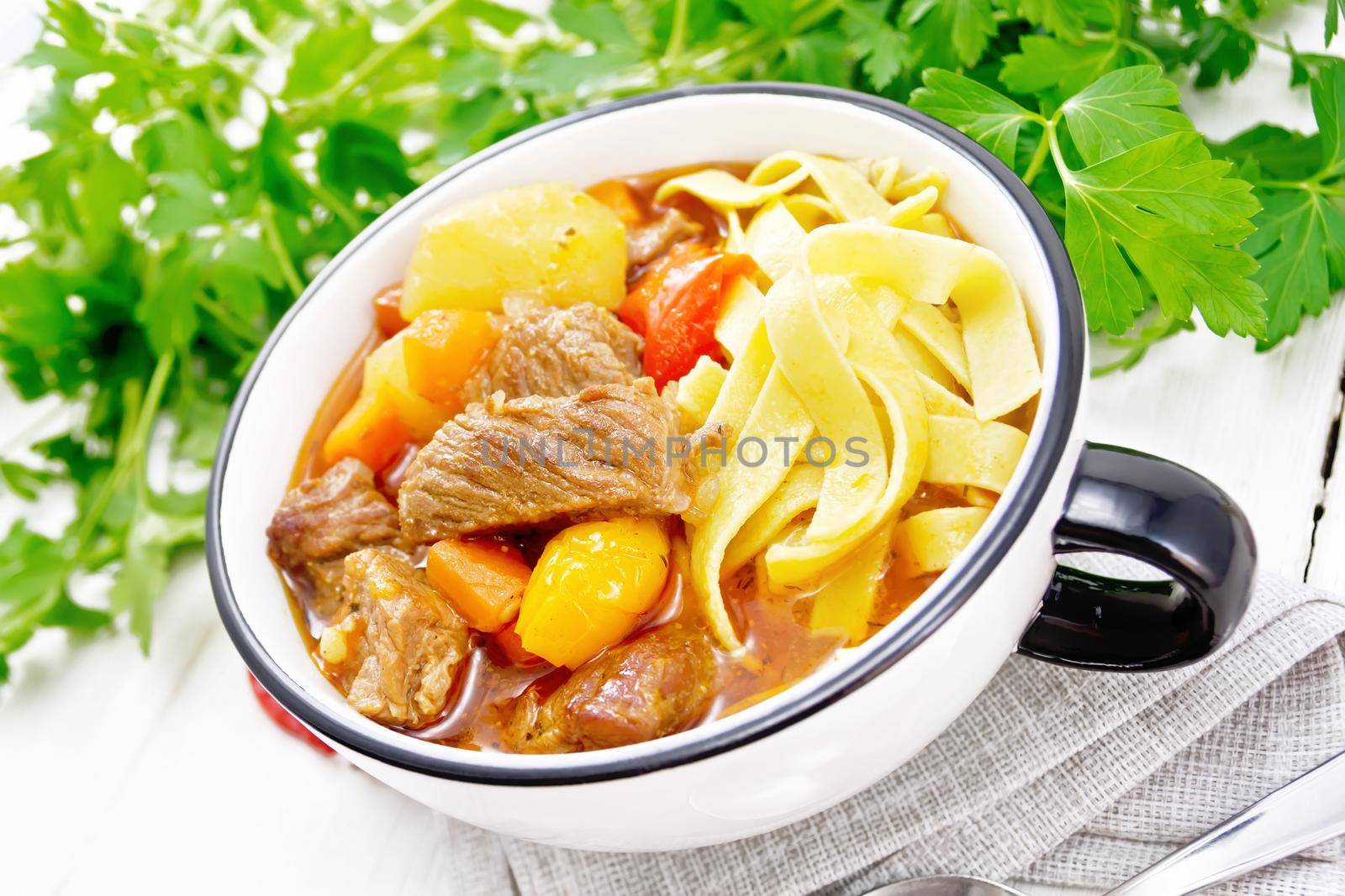 Central Asian national dish Lagman of meat, noodles and vegetables in bowl on a towel, garlic, parsley on background of light wooden board