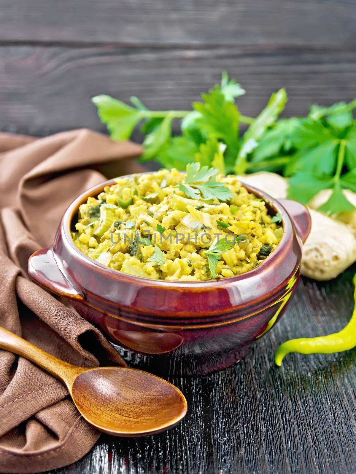 Indian national dish kichari made of mung bean, rice, celery, spinach, hot pepper and spices in a bowl on a towel, ginger and spoon on black wooden board background