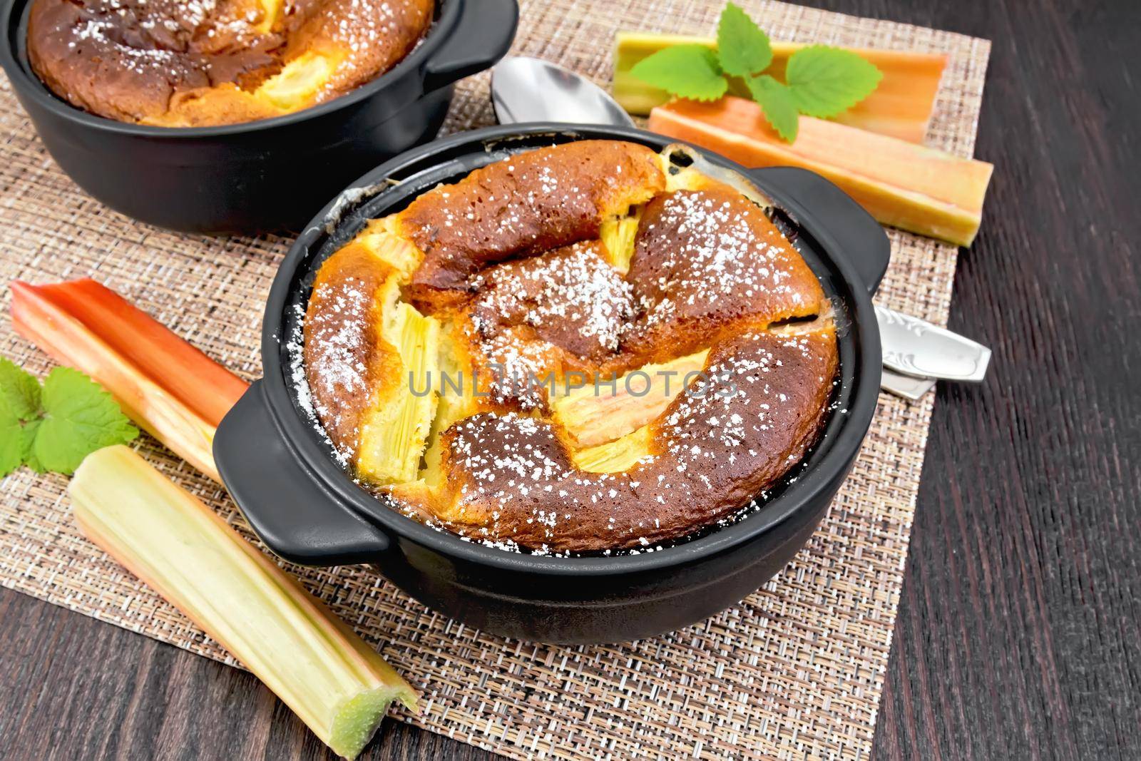 Pie clafouti with rhubarb in the brazier, mint and stalks of rhubarb on a napkin on the background of wooden board