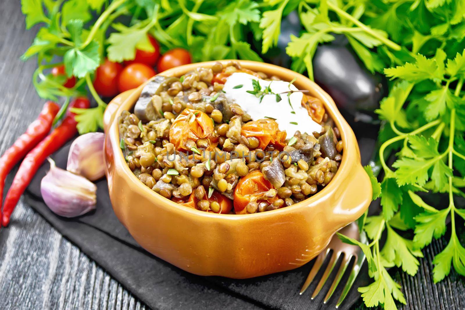 Green lentils stewed with eggplant, tomatoes, garlic and spices, sour cream sauce with a sprig of thyme in a bowl on a towel, parsley on wooden board background