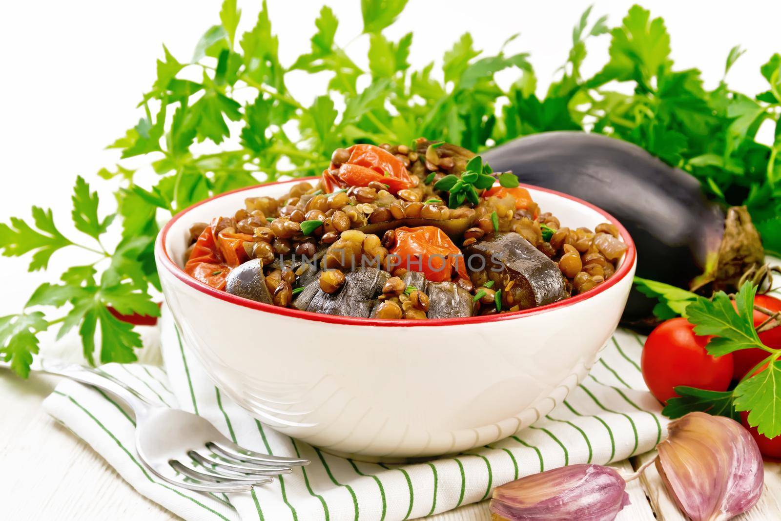 Green lentils stewed with eggplant, tomatoes, garlic and spices in a bowl on a towel, parsley on wooden board background