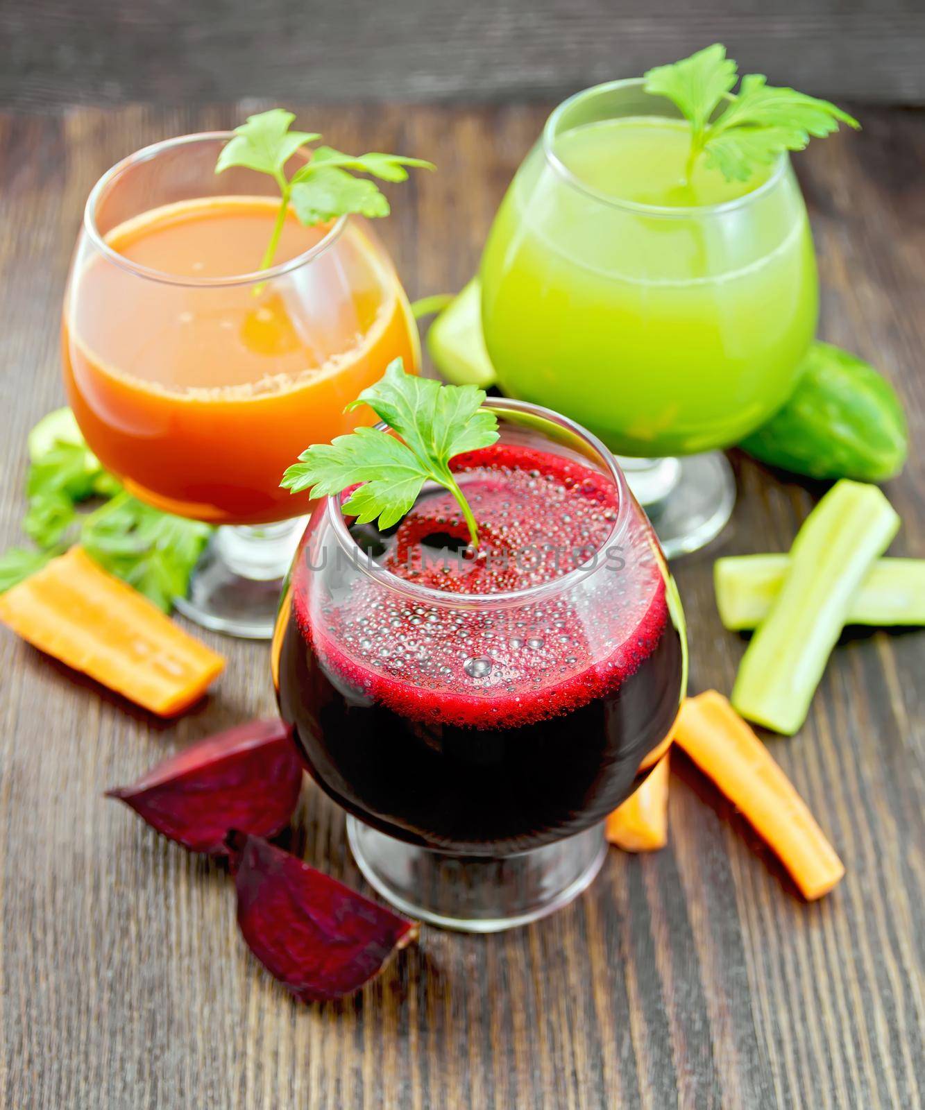 The juice of beet, carrot and cucumber in three wineglass, vegetables and parsley on a dark wooden board