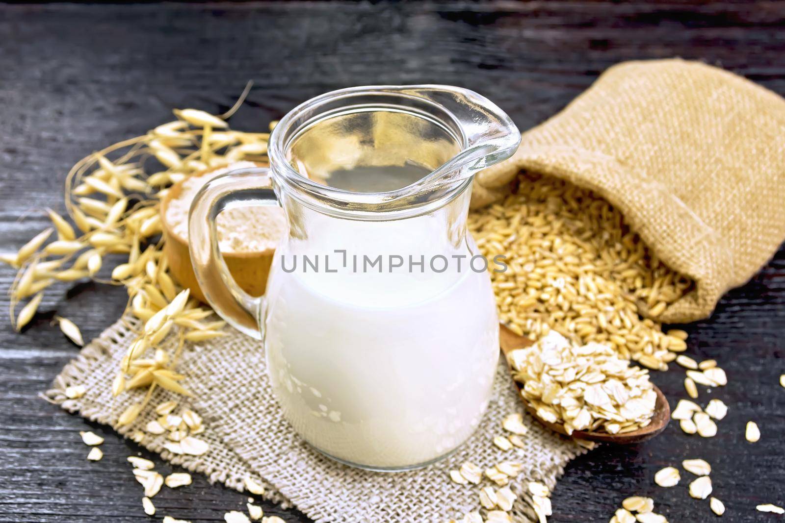 Oat milk in jug, flour in bowl, oatmeal in spoon, grain in jute bag, oaten stalks on burlap against dark wooden board