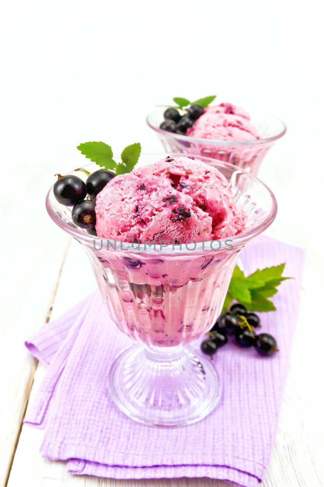 Ice cream with black currant in two glasses on a lilac napkin, berries with leaves against a light wooden board