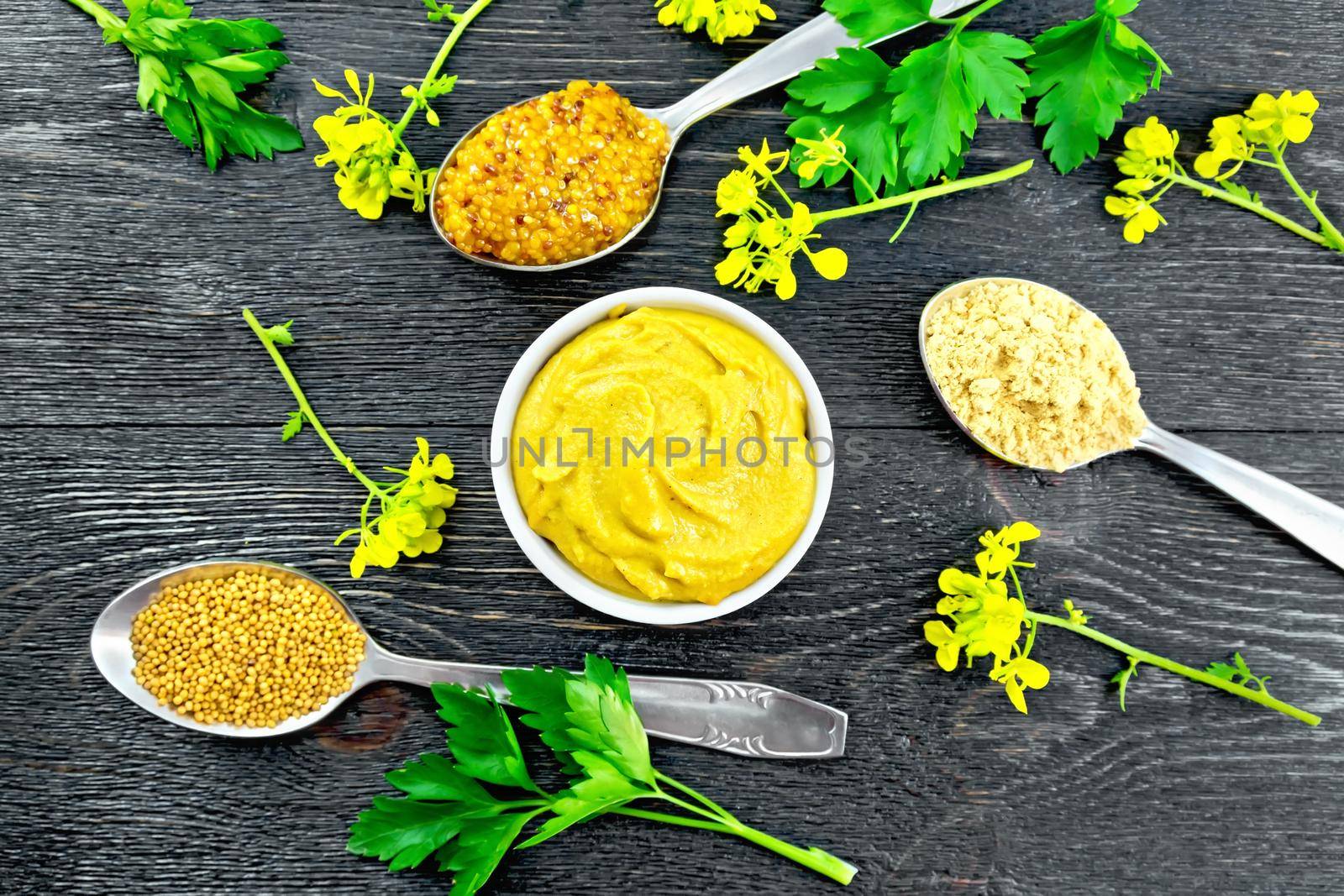 Mustard granular, powder, seeds in spoons and mustard sauce in a bowl, yellow flowers and parsley on the background of a black wooden board from above