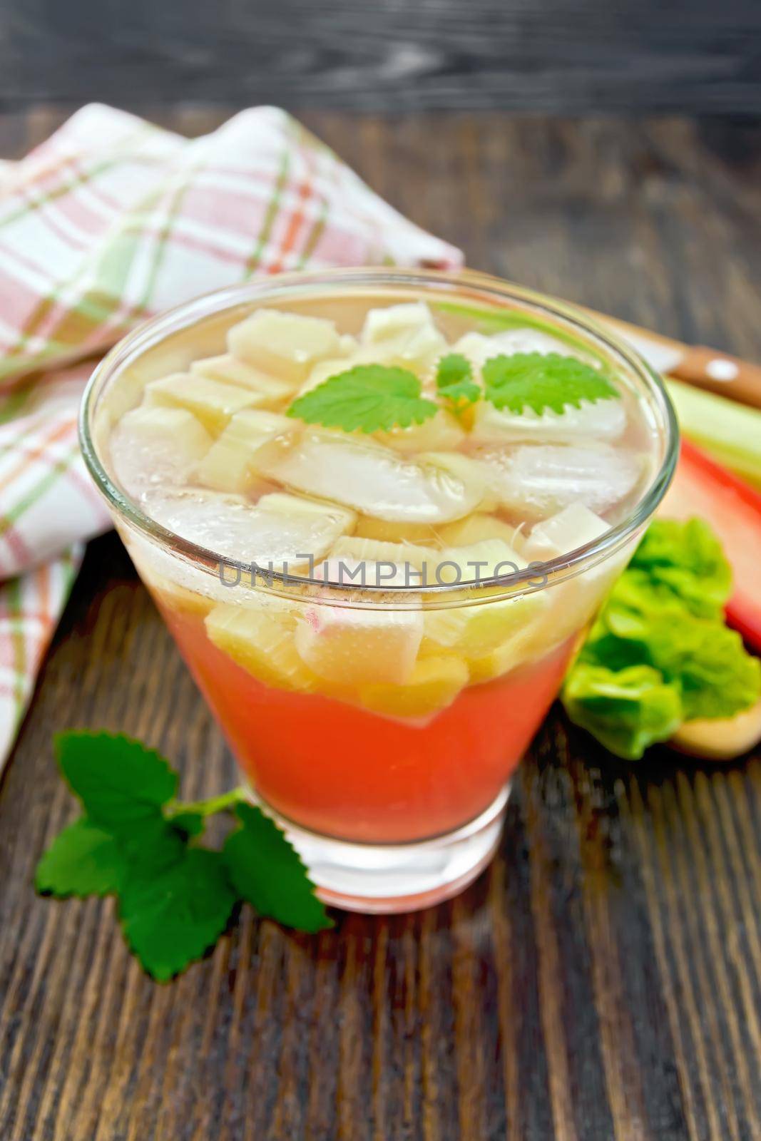 Lemonade with rhubarb and mint in a glass, stems and a leaf of a vegetable, napkin on a background of a dark wooden board
