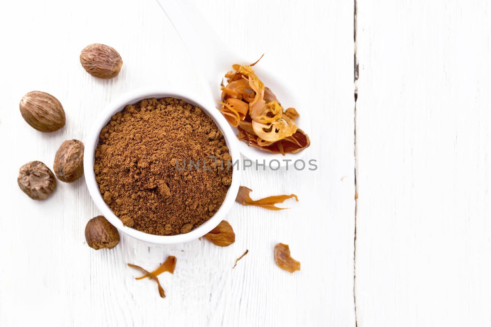 Ground nutmeg in a bowl and dried nutmeg arillus in a spoon, whole nuts on wooden board background from above