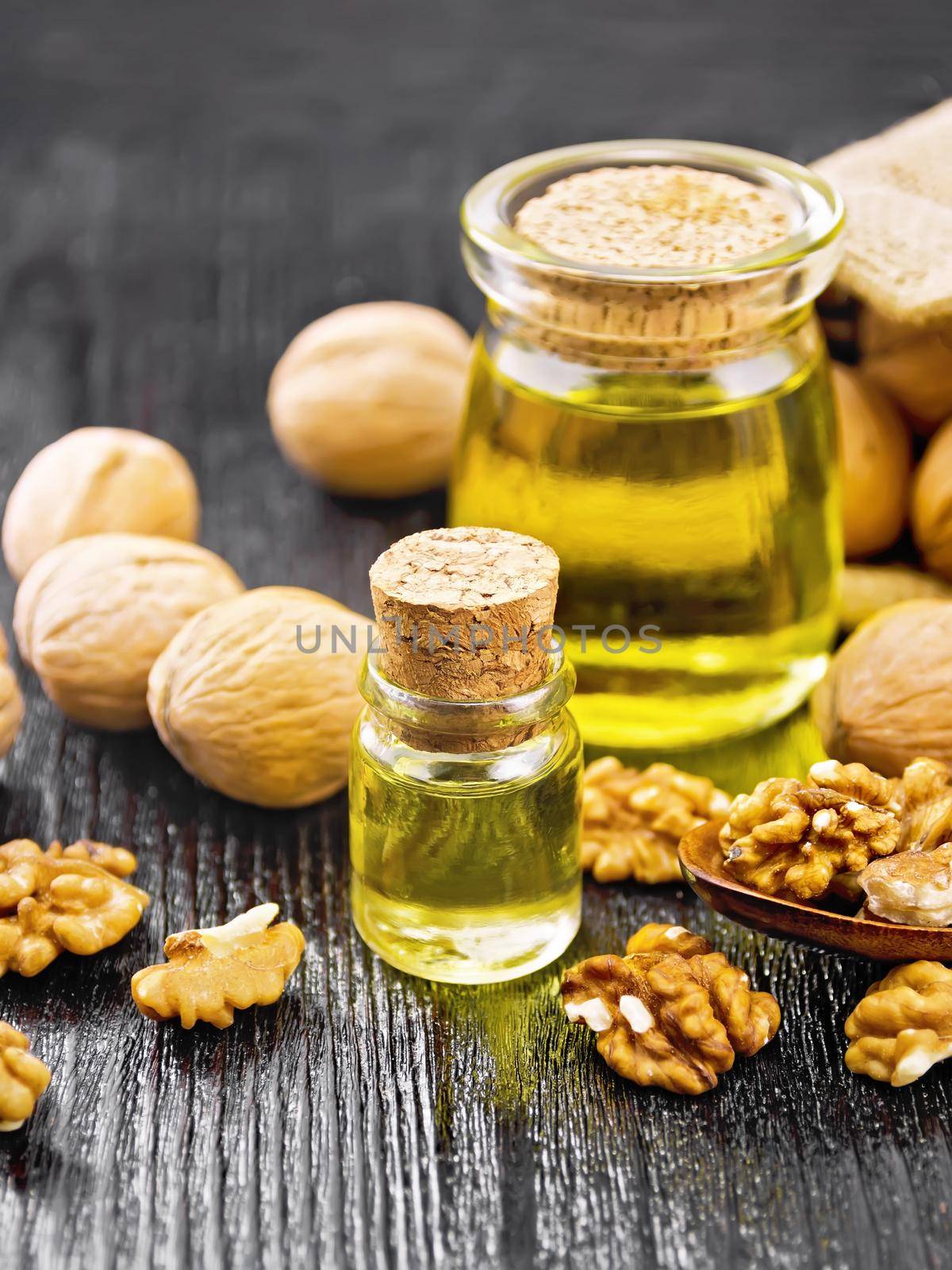 Walnut oil in a glass bottle and a jar, nuts in bag, spoon and on table against black wooden board