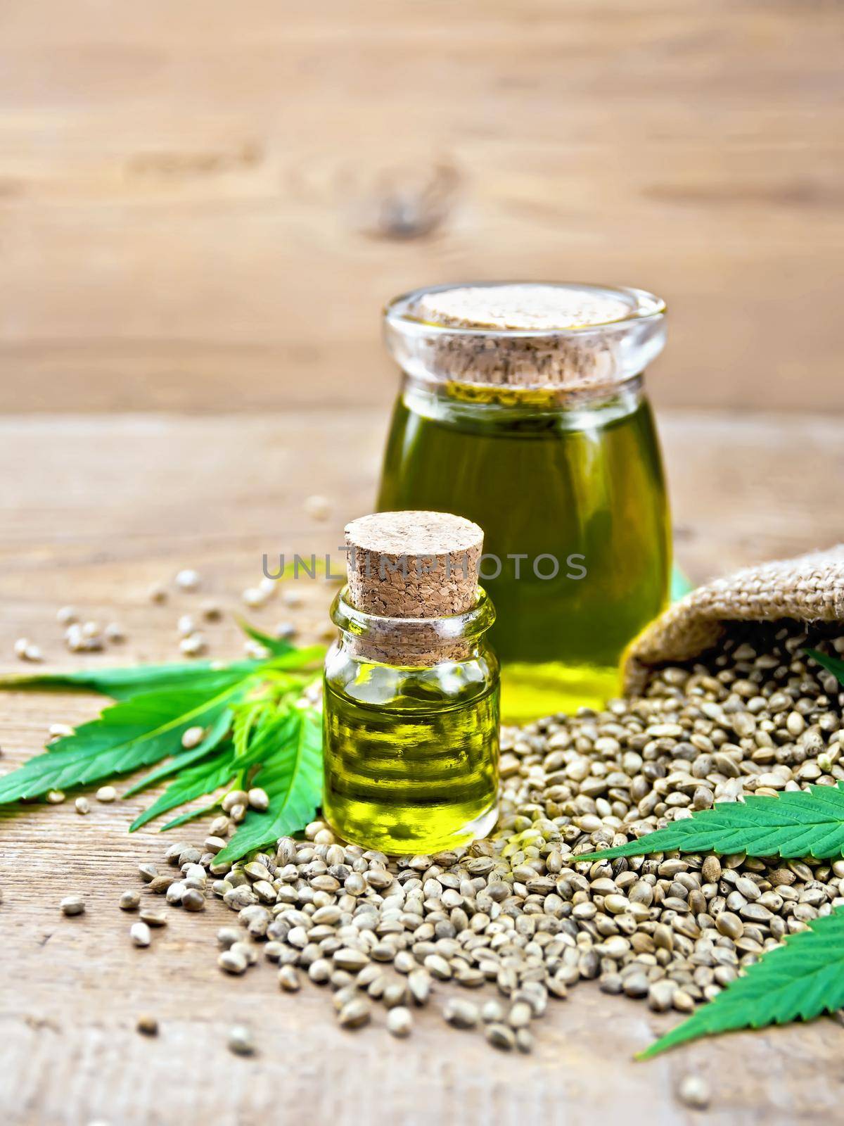 Hemp oil in two glass jars with grain in the sack, leaves and stalks of cannabis on the background of wooden boards
