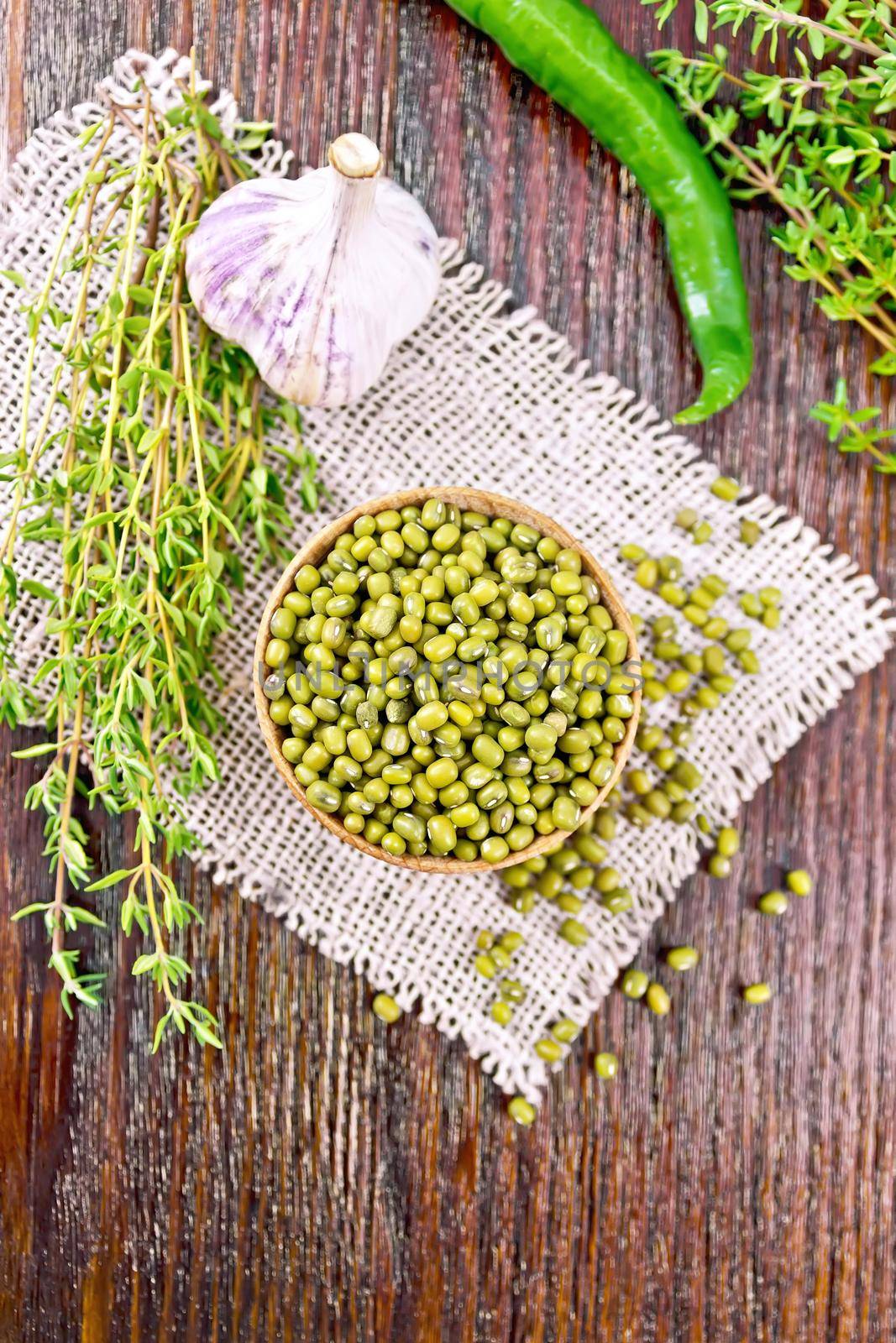 Green lentils mung in a wooden bowl with thyme and garlic on sackcloth, hot peppers on a wooden board background from above