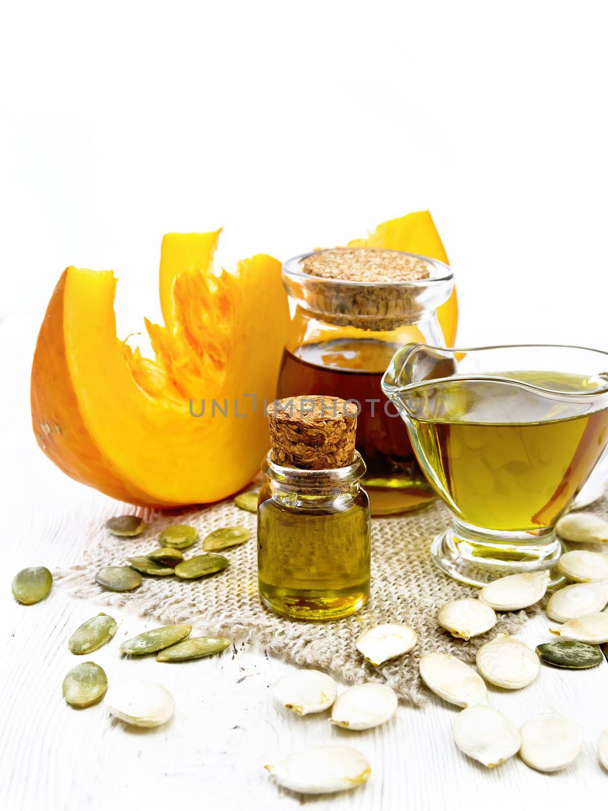 Pumpkin oil in a vial, gravy boat and a jar on burlap, seeds on the table, slices of vegetable on light wooden board background
