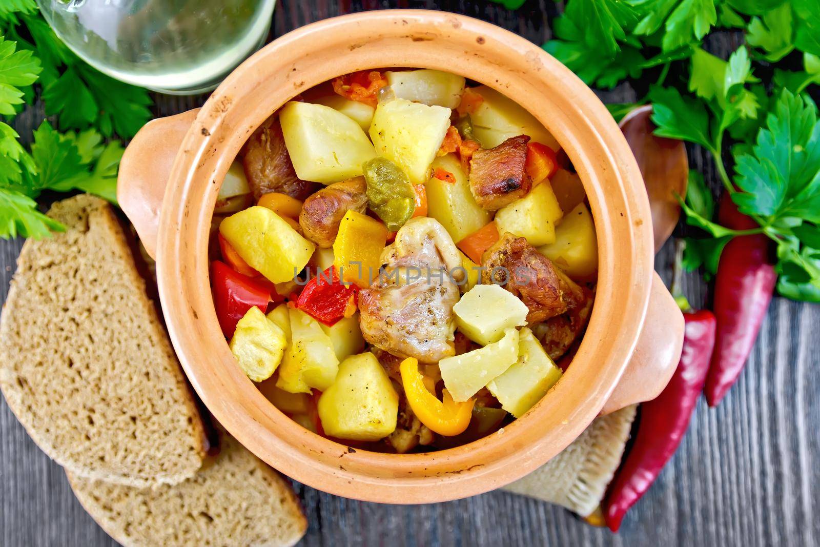 Roast with chicken, potatoes, squash and sweet peppers in a clay pot on a napkin of burlap, bread and parsley on a background of wooden boards on top
