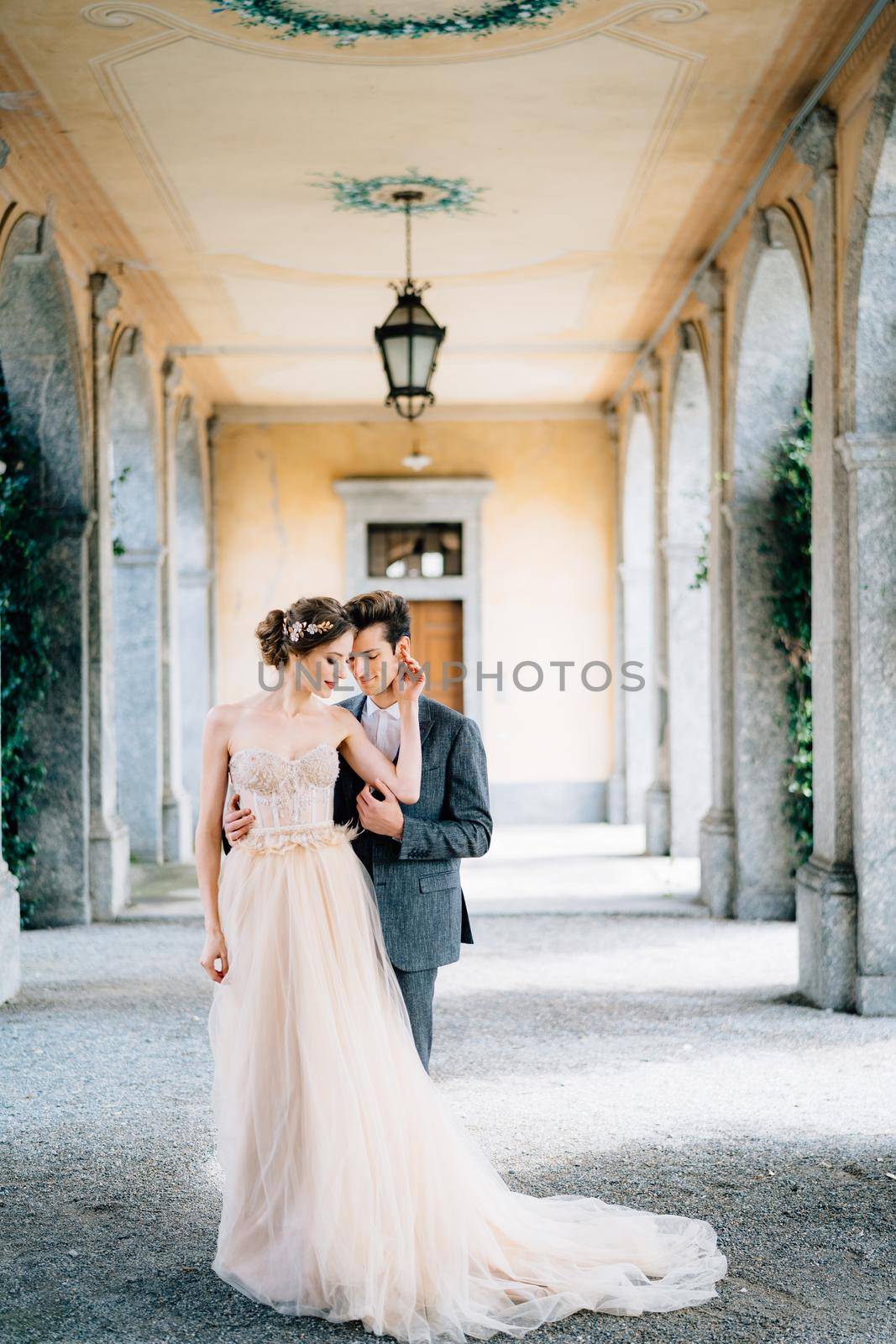 Smiling groom hugs bride on an old terrace with columns entwined with green ivy. Lake Como, Italy. High quality photo