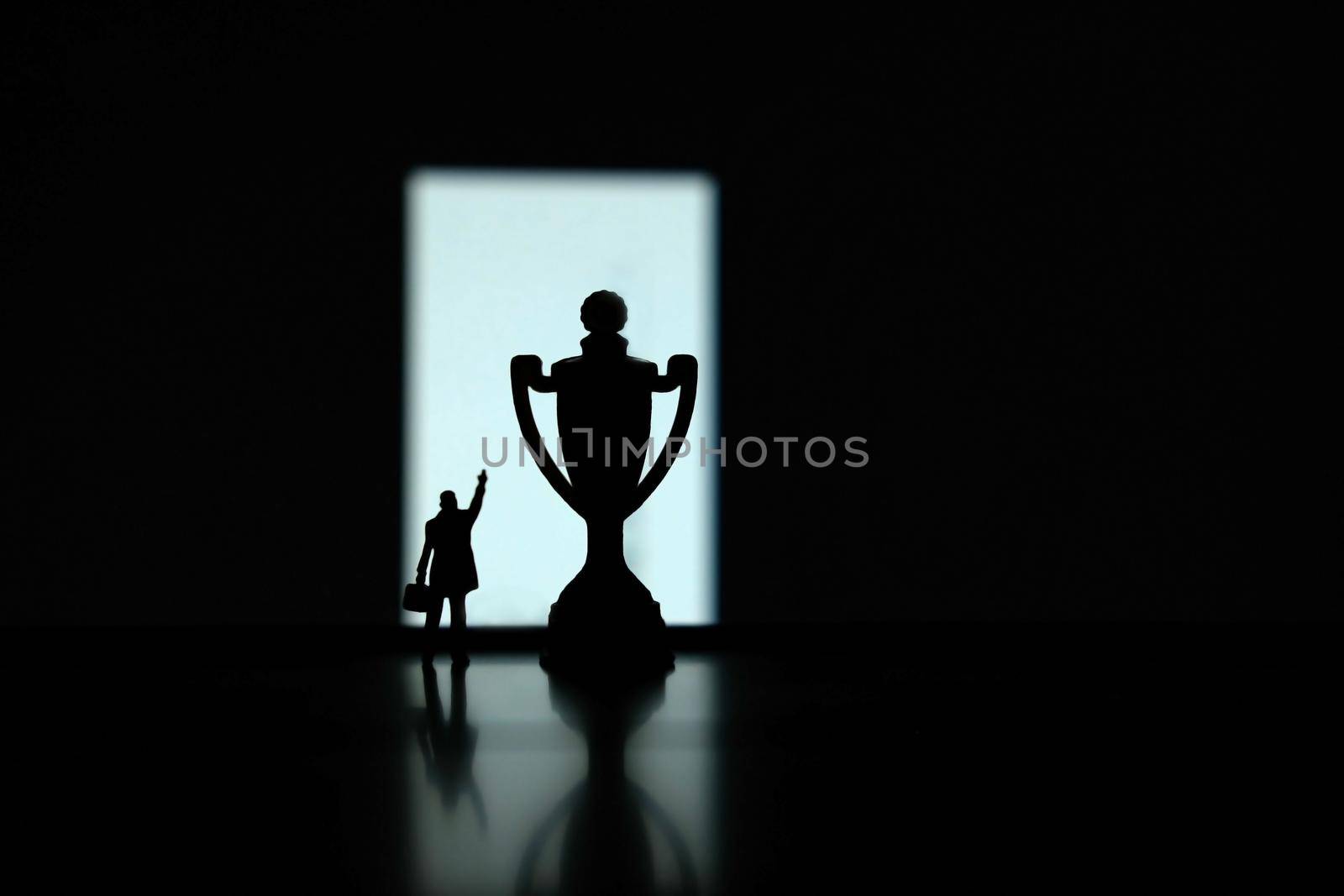 Business strategy conceptual photo - Silhouette of miniature businessman pointing on winning trophy in the podium by Macrostud