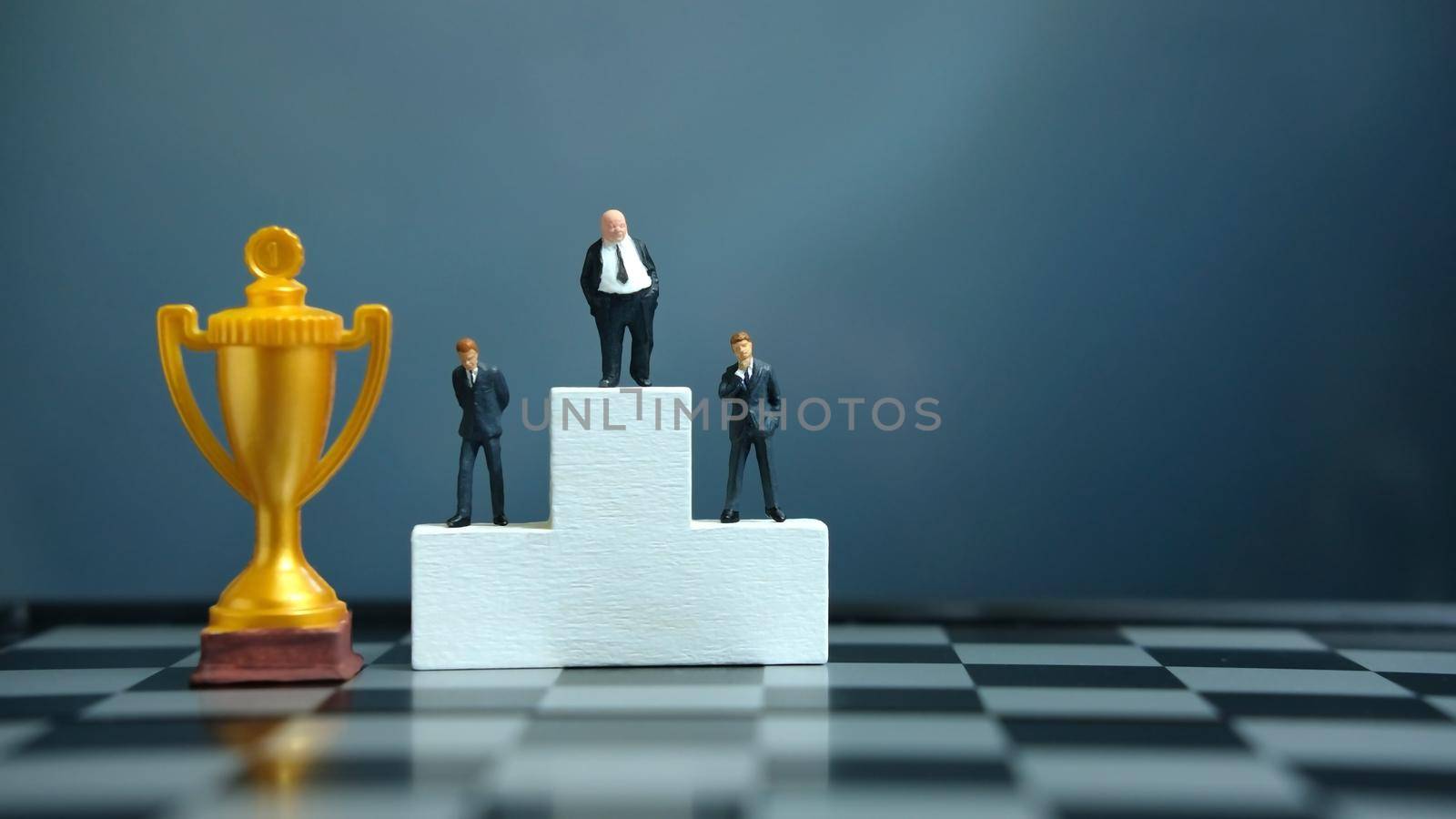Miniature business concept - businessman standing on white winner podium with golden and silver trophy. Image photo