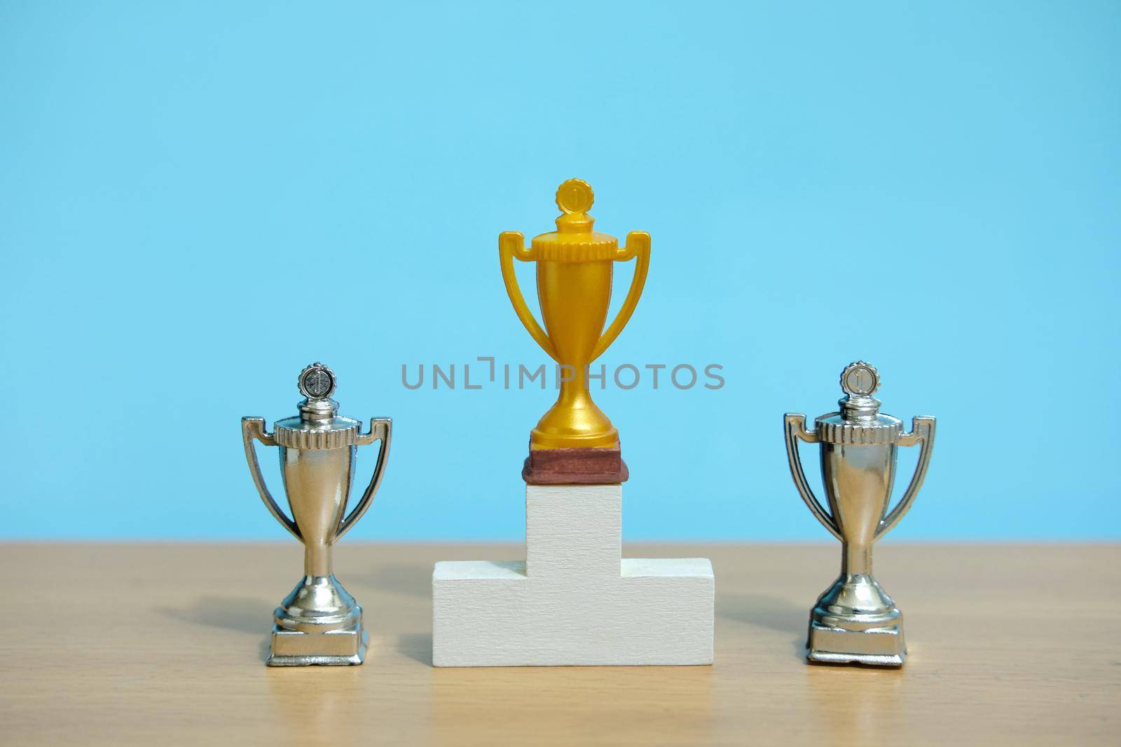 Golden trophy standing on white podium with two silver trophy at back by Macrostud