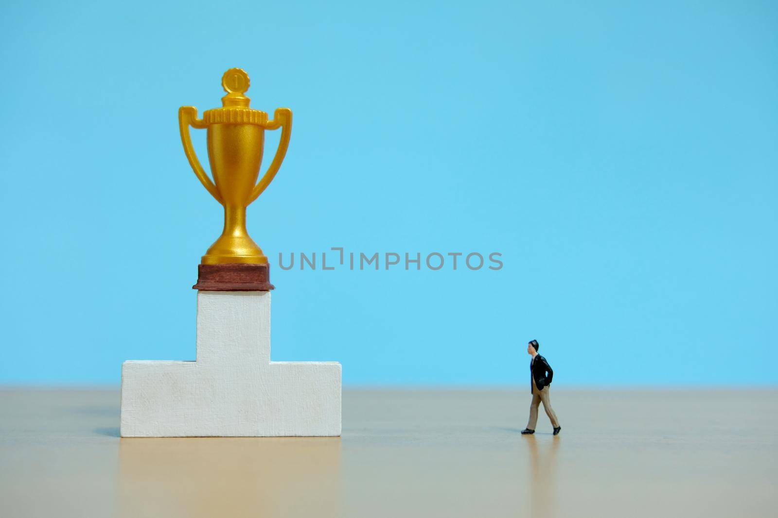 Miniature business concept - businessman walking toward golden trophy above white podium. Image photo