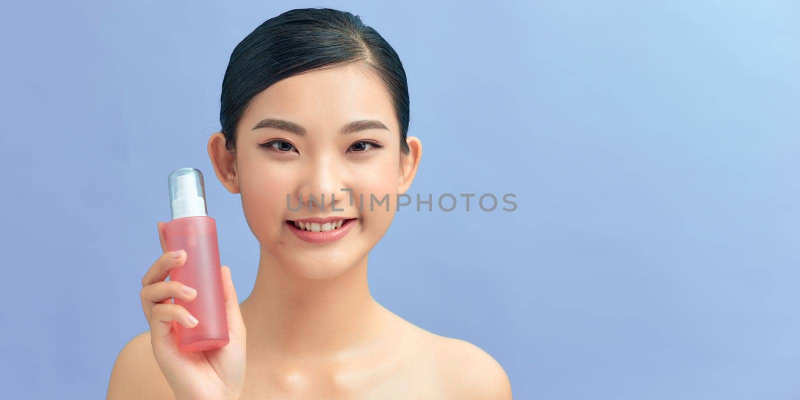 Smiling young girl demonstrates moisturizing tonic in pink bottle. by makidotvn
