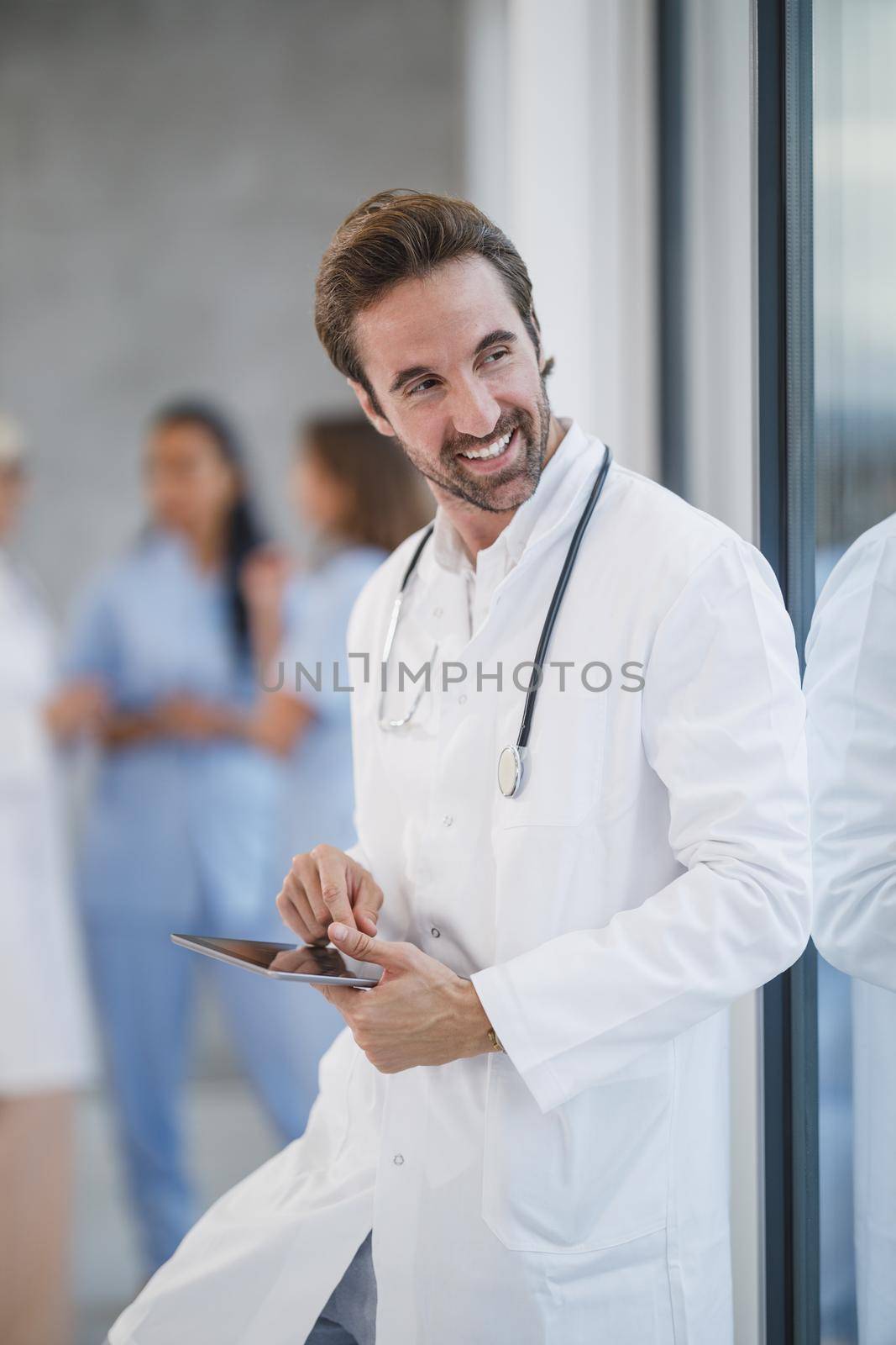 Shot of a smiling doctor using digital tablet and pensive looking out the window while having quick break in a hospital hallway.