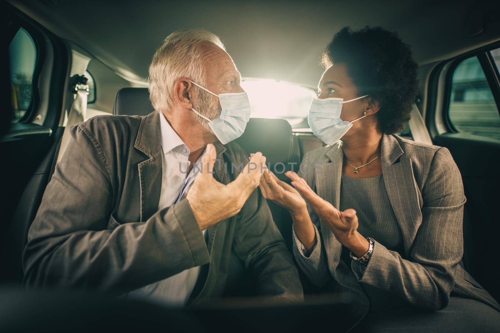 Shot of two worried multi-ethnic business people with protective mask having a discussion about work while sitting in the backseat of a car on their morning commute during COVID-19 pandemic.