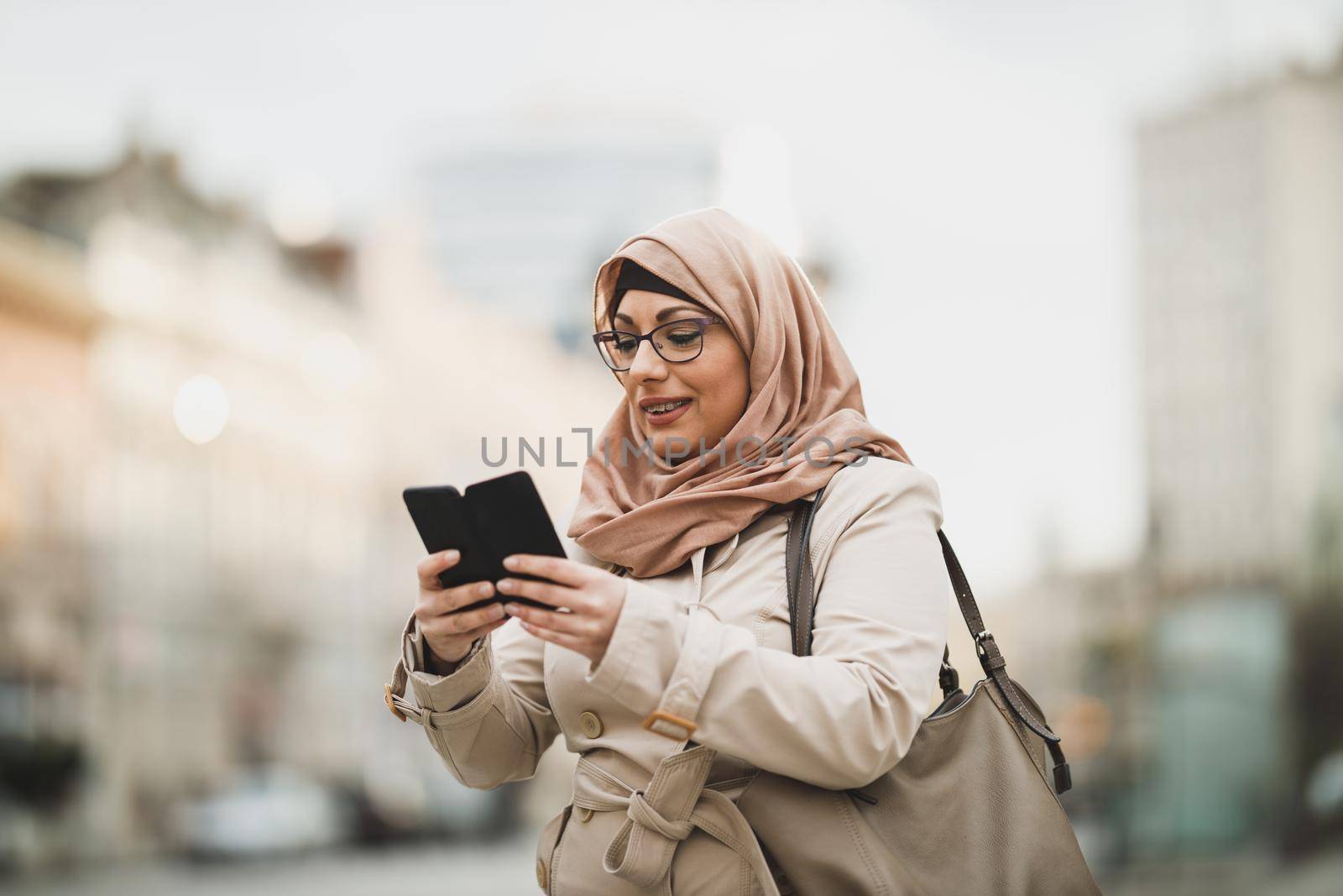 Smiling Muslim woman wearing a hijab and tipping messages on her smartphone while standing in urban environment.